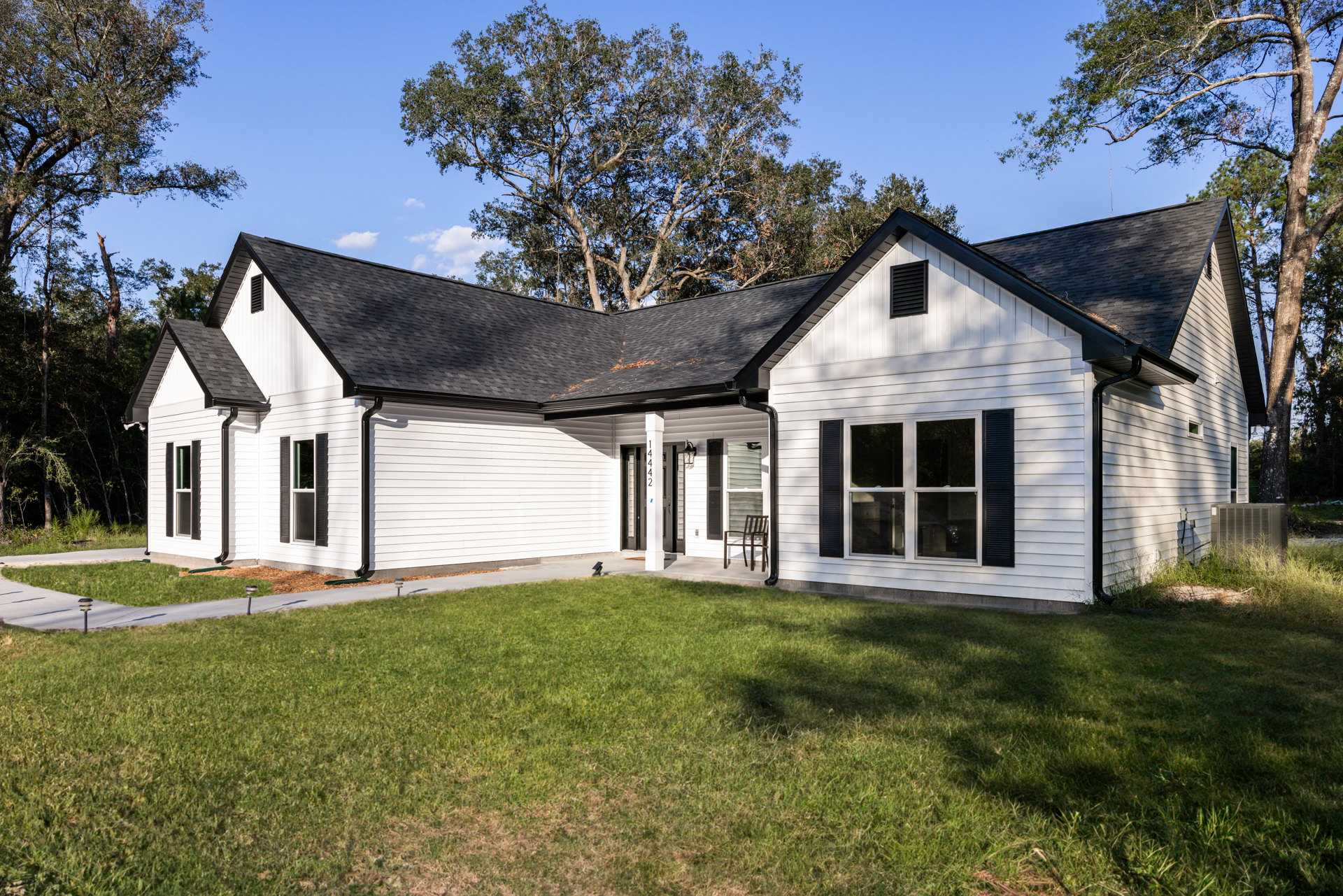 White house with black roof, glass-paned windows, black vent, front porch with chair, surrounded by green lawn and trees under blue sky