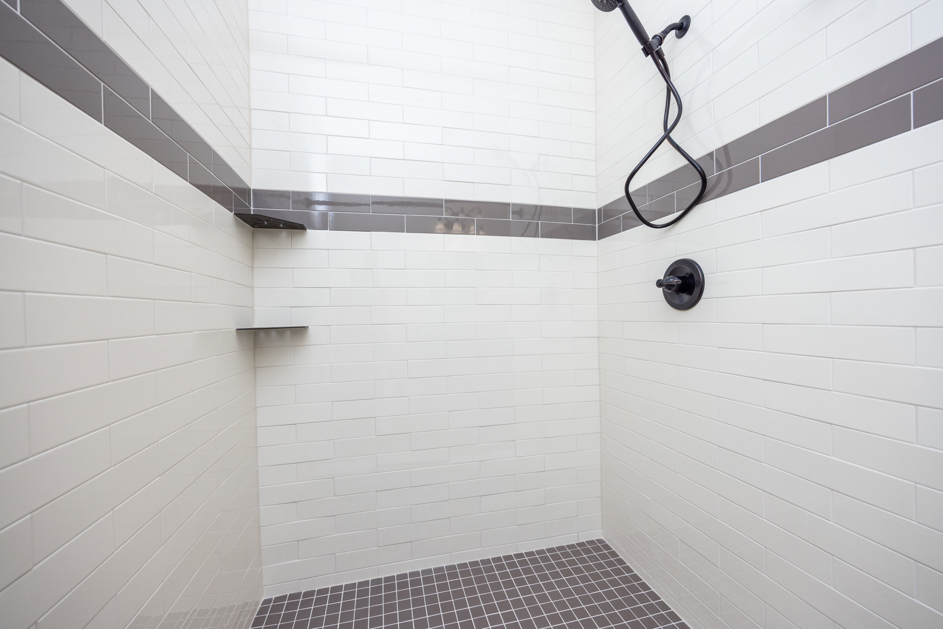 White tile shower with dual shower heads, grey accent tiles, black hose and pipe, close-up of wall switch, tiled floor