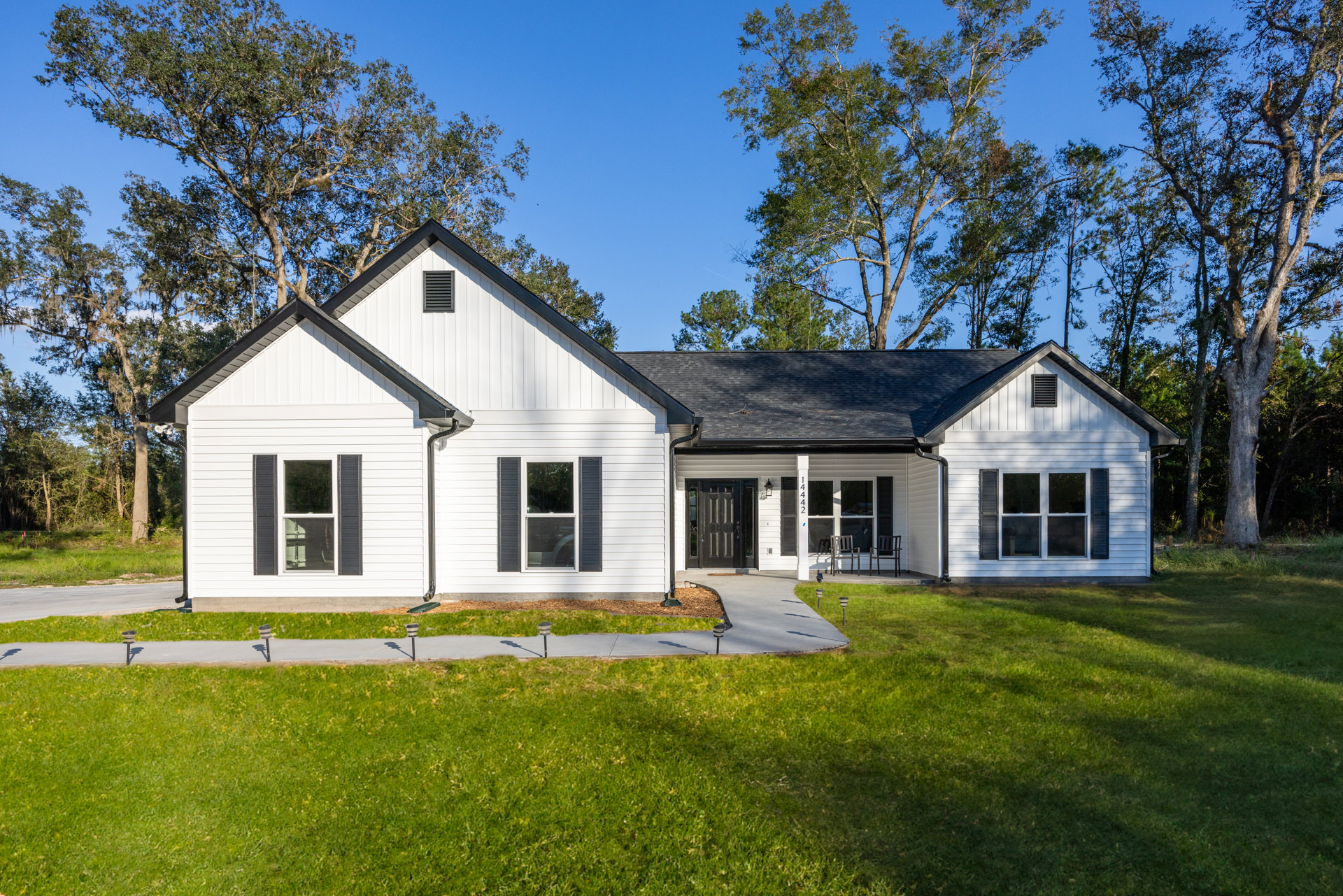 White house with black shutters, white-framed windows, and a covered porch, surrounded by green lawn and mature trees in the background