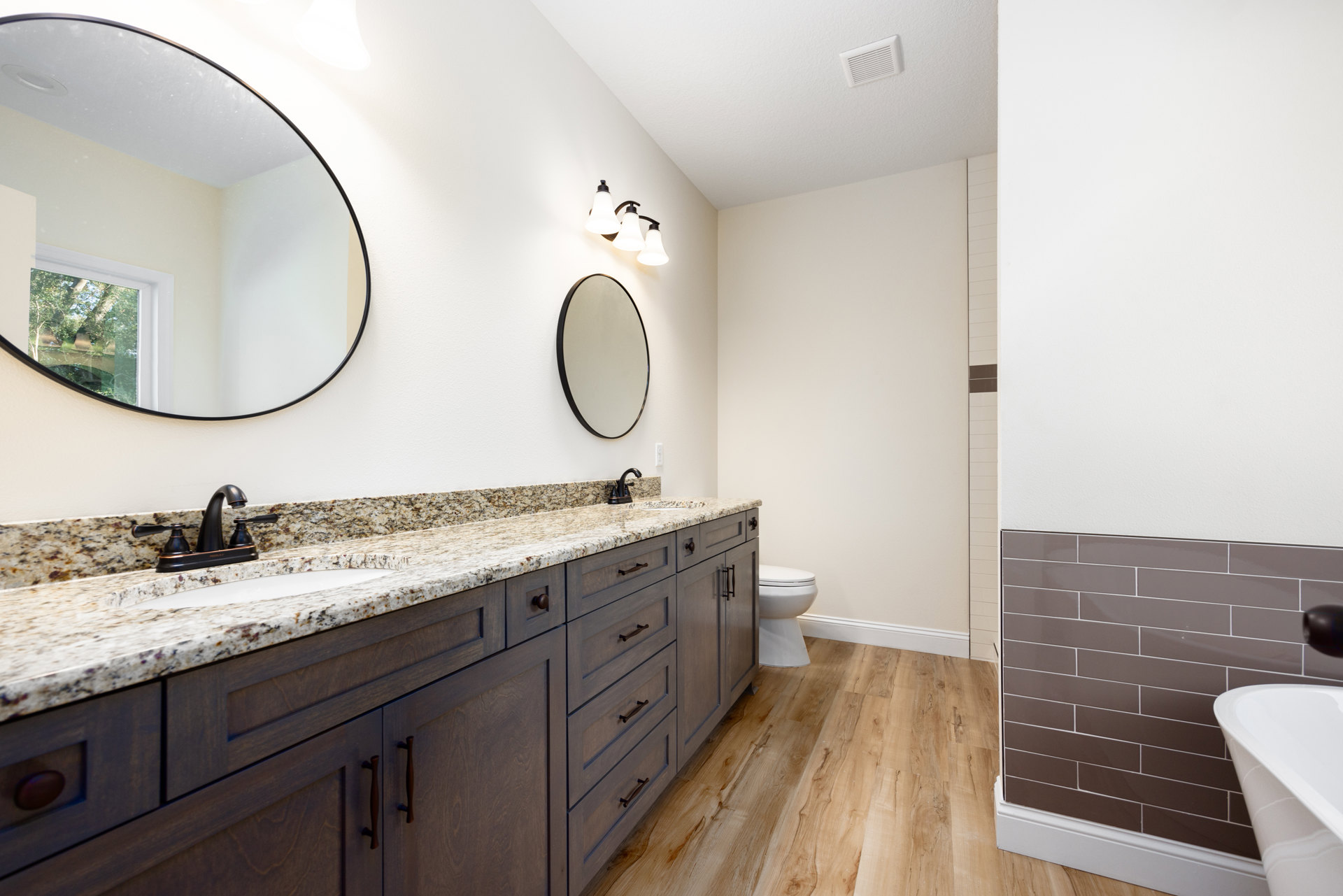 White bathroom with round black-framed mirror above rectangular sink, light gray tile backsplash, white countertop, chrome faucet, closed toilet, and white wall vent.