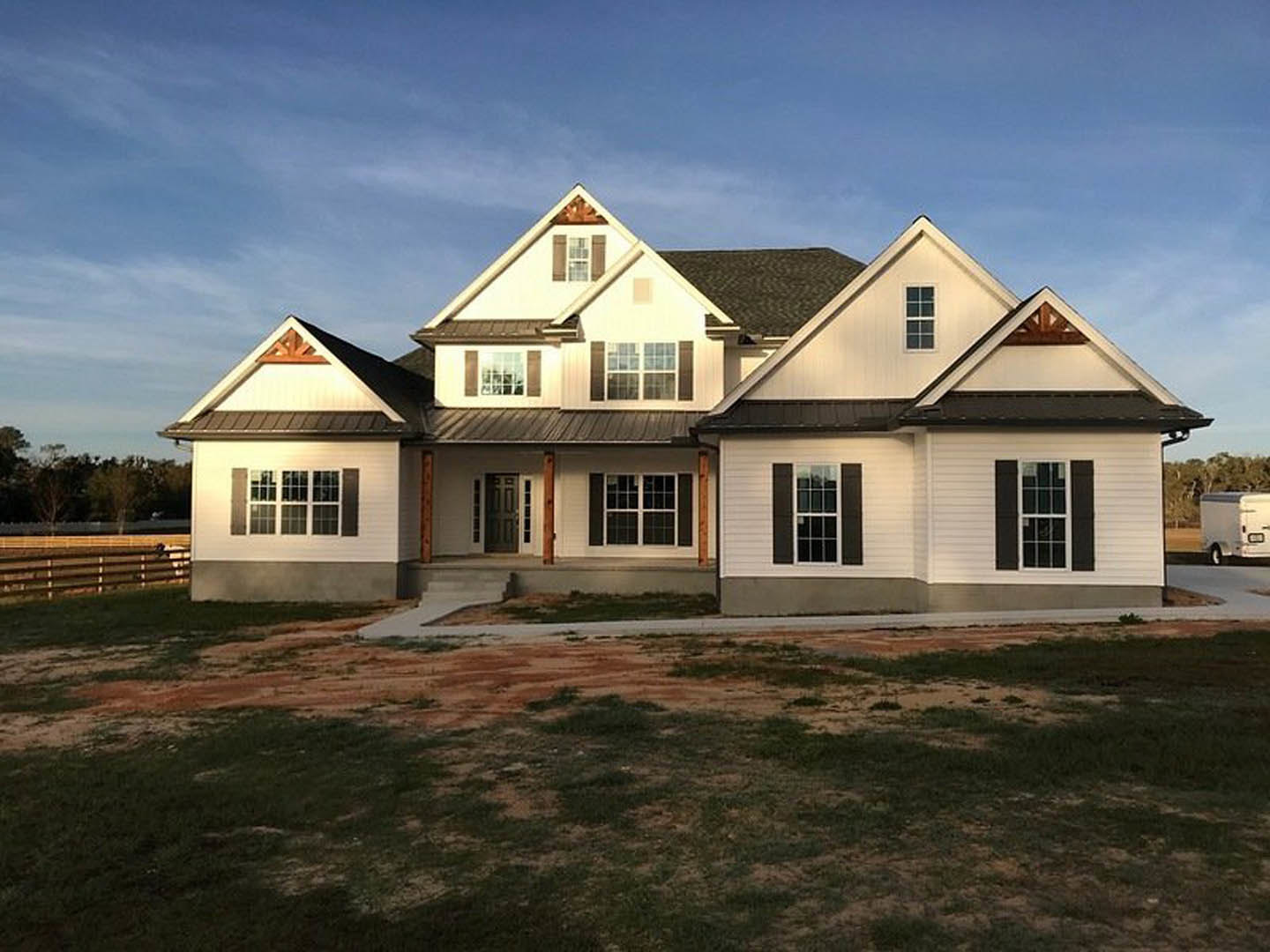 White two-story house with black roof, covered front porch, multiple white-framed windows, expansive grassy yard, and a white trailer parked on the driveway.
