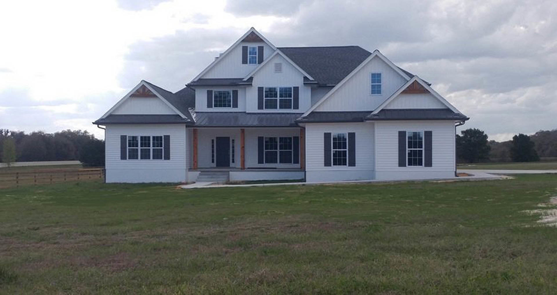 White two-story house with grey roof, multiple pane windows, covered porch, and expansive green lawn under partly cloudy sky
