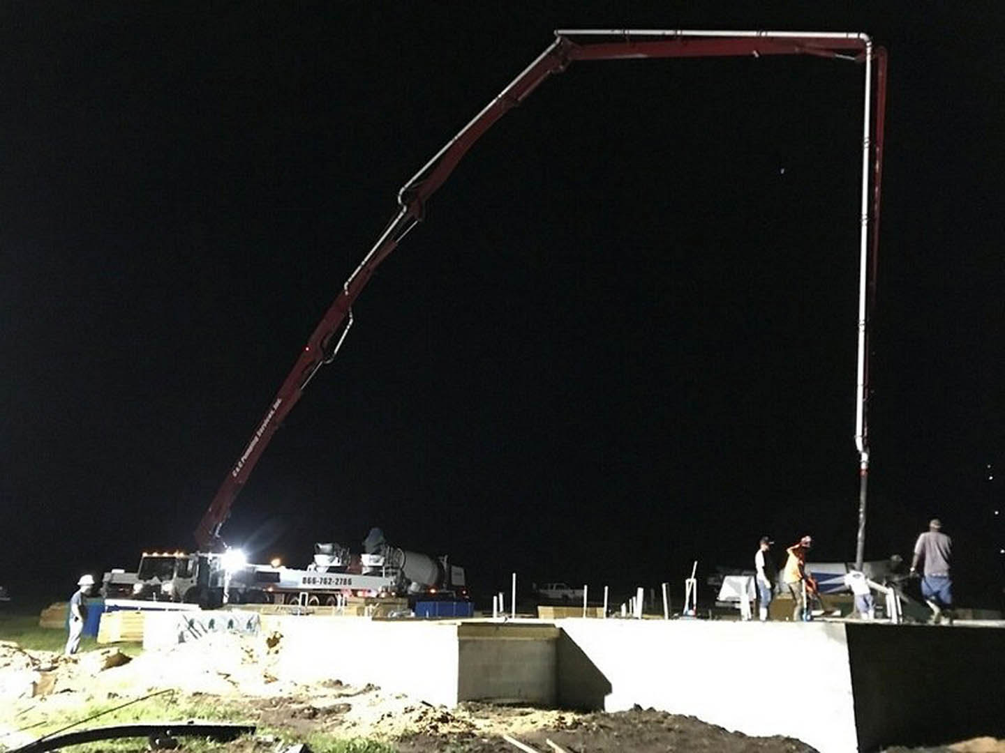A crane hoisting a concrete slab at night outside a custom home, with construction lights illuminating the scene and a white vehicle parked nearby.
