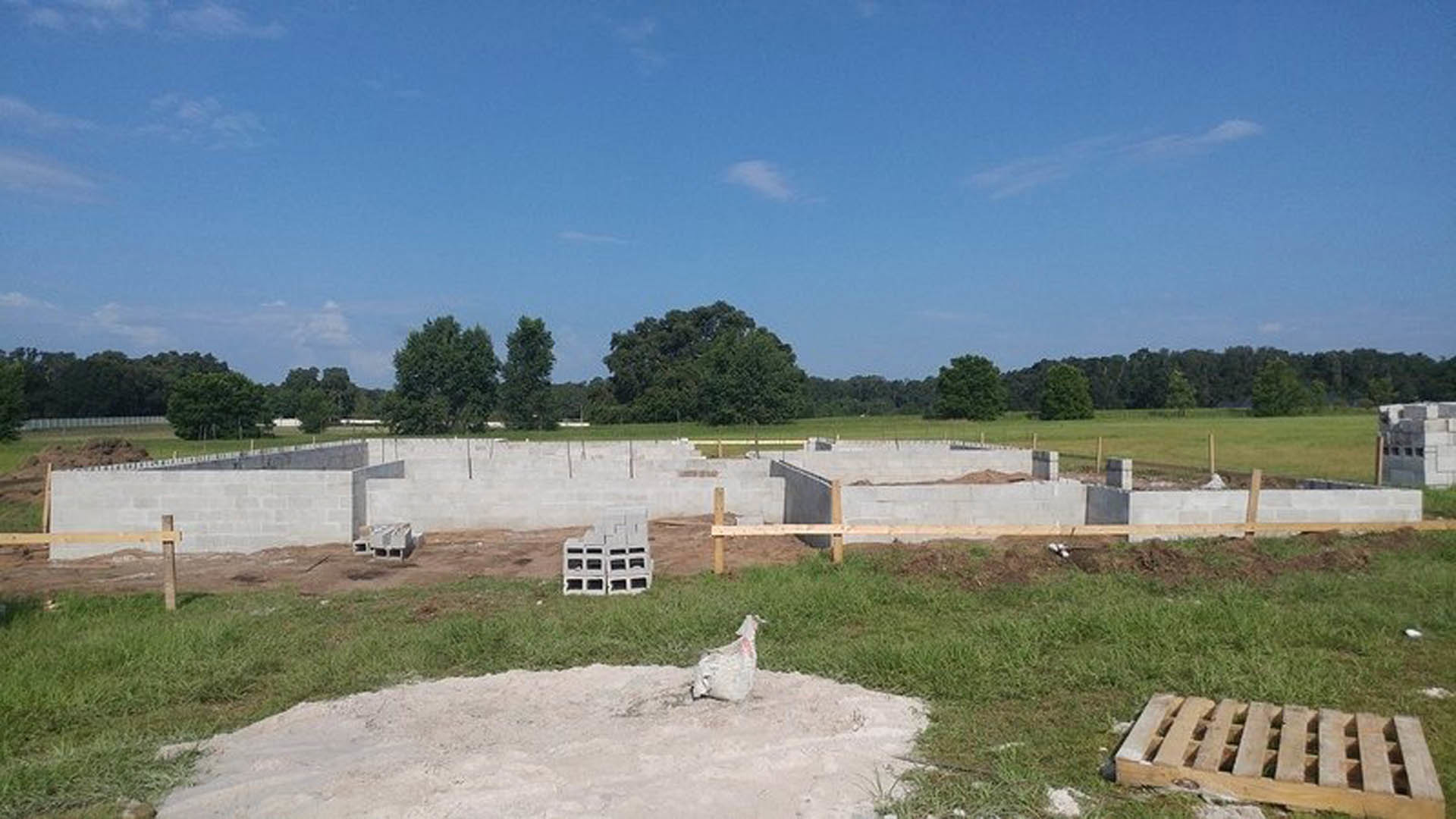 Concrete foundation surrounded by grass and dirt, wooden pallet and stack of white blocks nearby, sheep grazing in the background, blue sky with scattered clouds overhead.