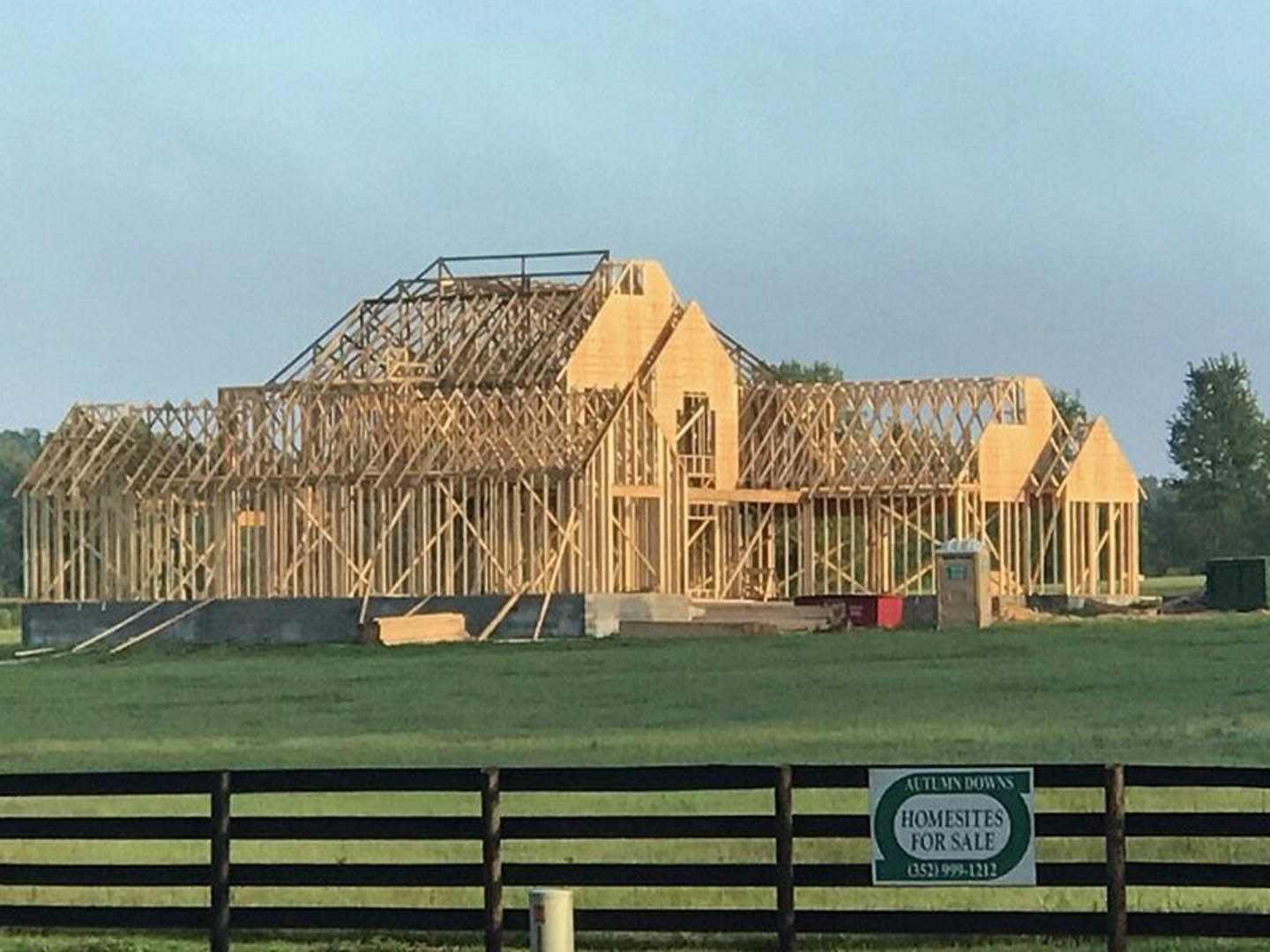 Wood-framed house under construction on grassy field, blue sky overhead, tree with green leaves nearby, temporary fencing and construction sign visible