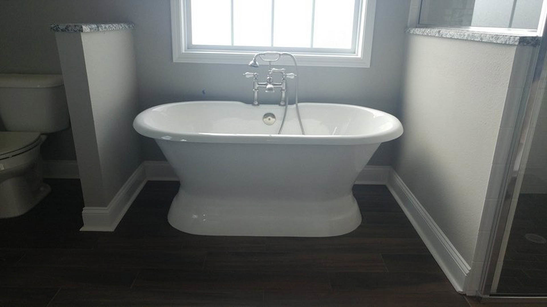 Freestanding white bathtub with chrome faucet beside large window, gray tile flooring, and partial view of toilet in modern bathroom
