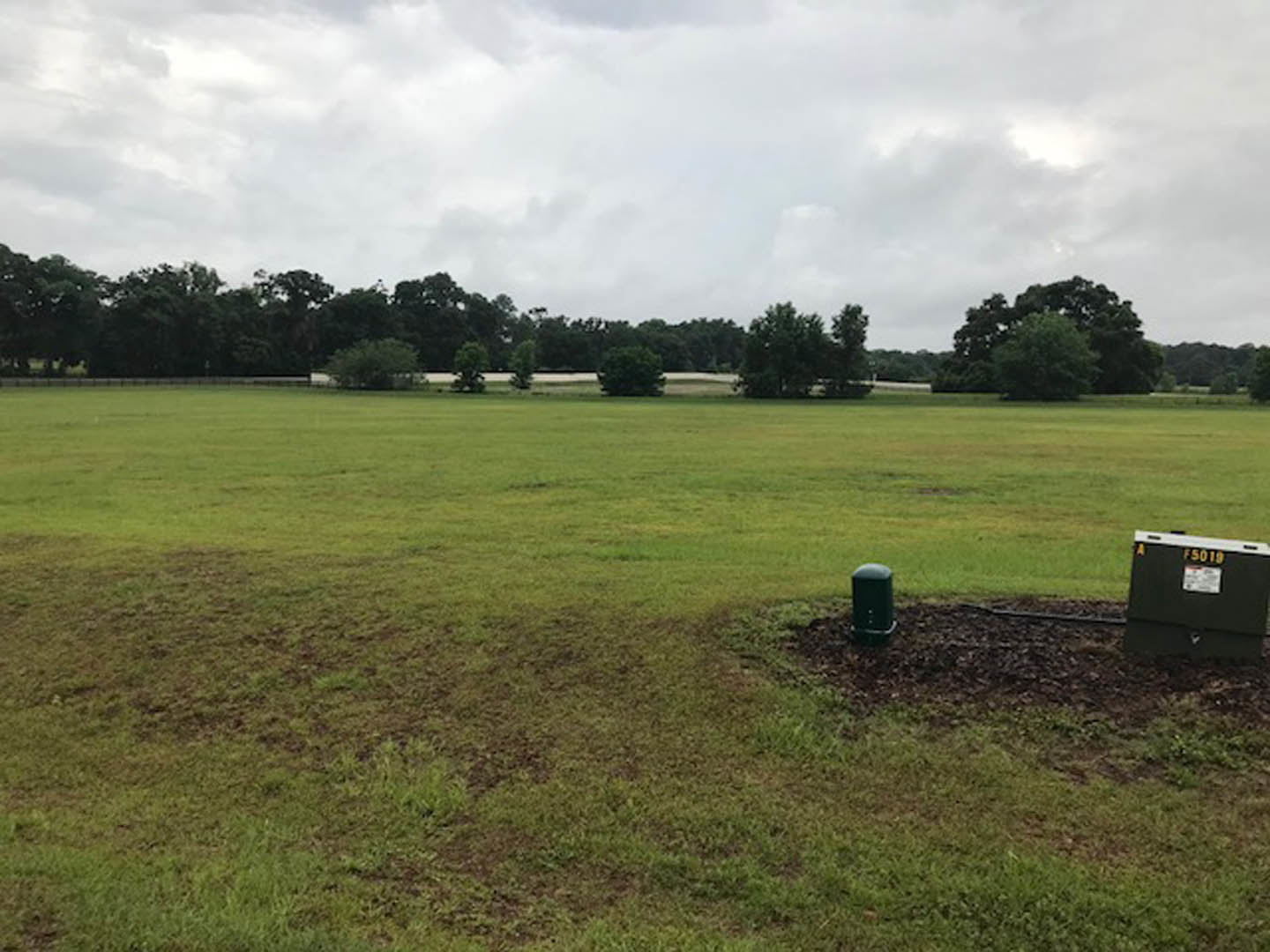 Green grassy field with scattered trees under a cloudy sky, black waste container with yellow numbers mounted on a green pole in the foreground.
