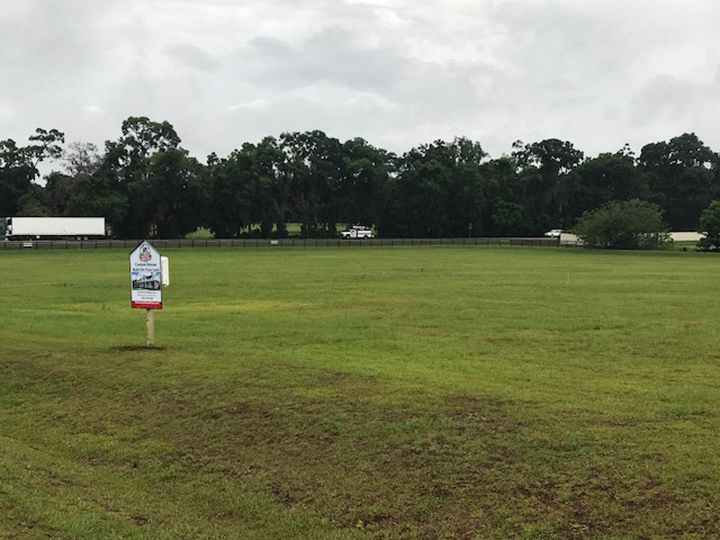 Grassy field with scattered trees under cloudy sky, white rectangular sign with black text placed near lawn