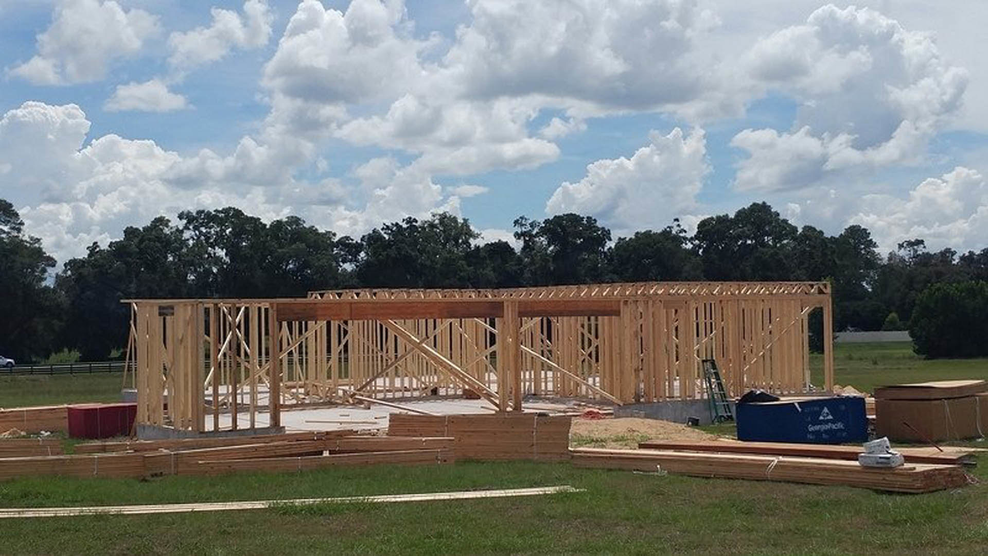 Wood-framed custom home under construction with exposed beams, surrounded by grass and trees, cloudy sky overhead, blue utility box and hot tub visible in foreground