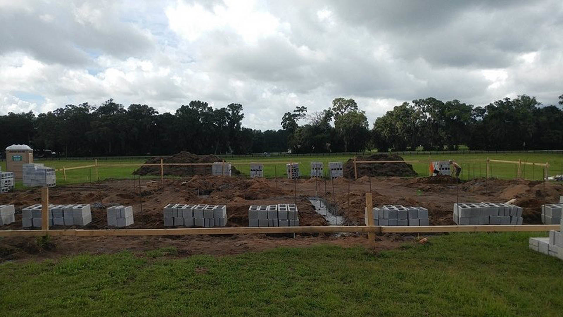 Cement blocks stacked on dirt at a construction site, surrounded by grass, trees, and a cloudy sky