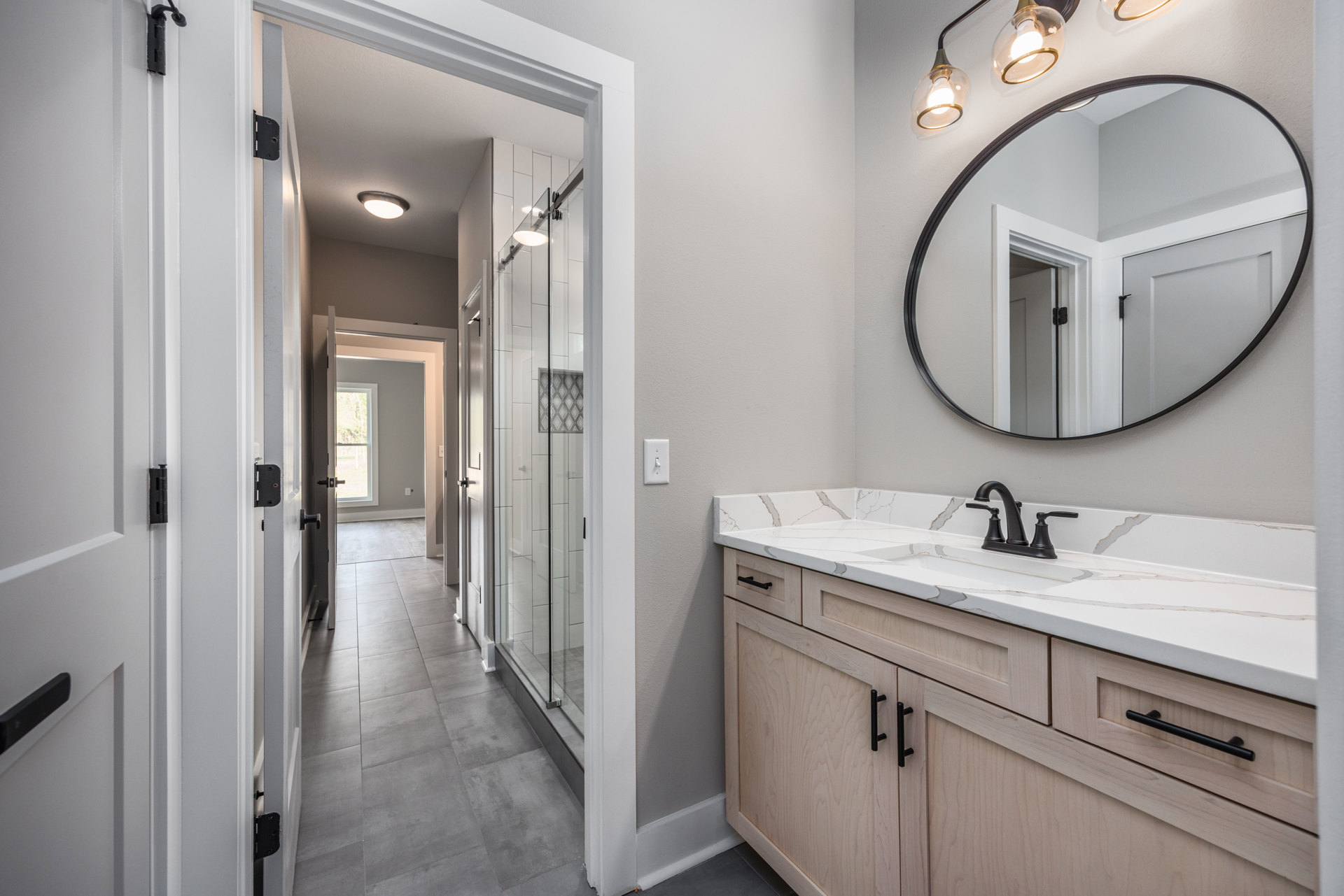 Bathroom with a round wall mirror above a white countertop, black faucet, wooden vanity with black metal bar, walk-in shower with tiled walls, and two-bulb light fixture