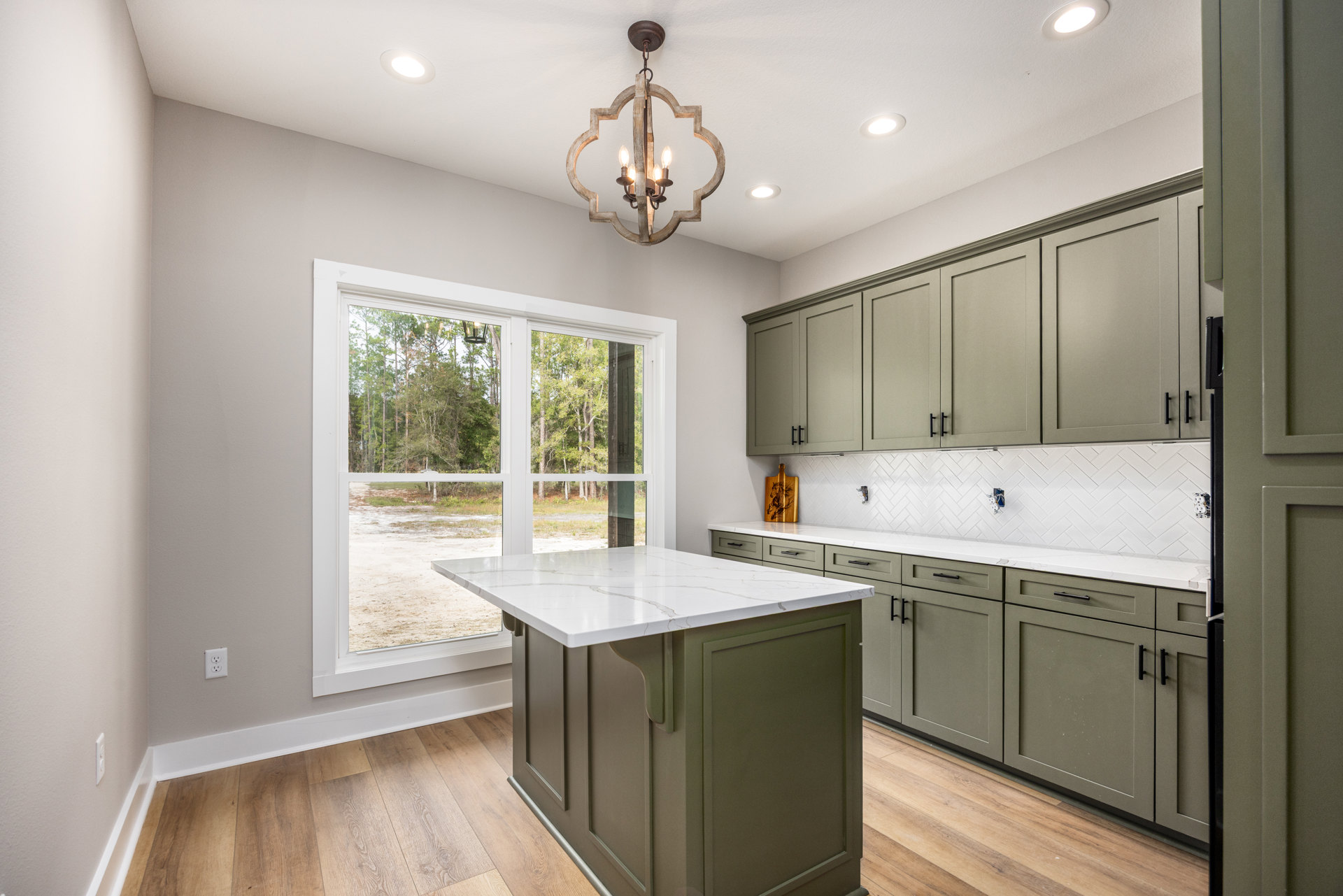 Spacious kitchen featuring a large white marble island with brown trim, grey cabinetry, tile flooring, stainless steel sink, and a wooden chandelier with three lights; window above