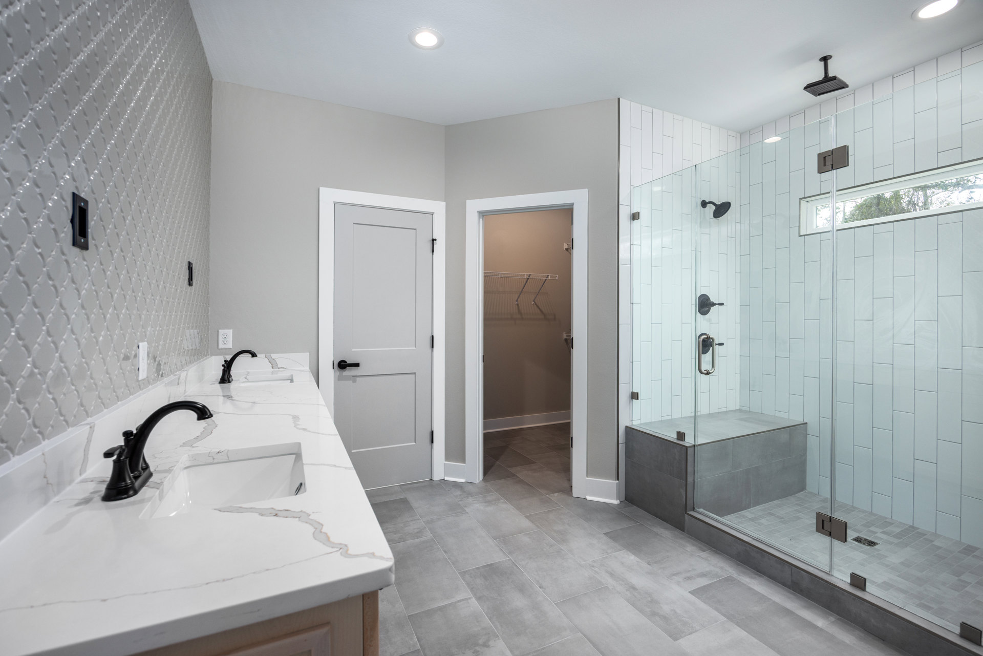 Bathroom with double sinks on a white marble countertop, black faucets and hardware, glass-enclosed shower with ceiling-mounted black showerhead, white door with black handle