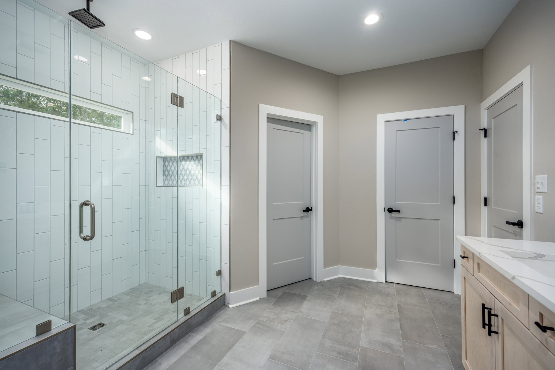Bathroom with frameless glass shower enclosure, white door featuring black handle, ceiling light fixture, tiled walls and floor, chrome shower hardware