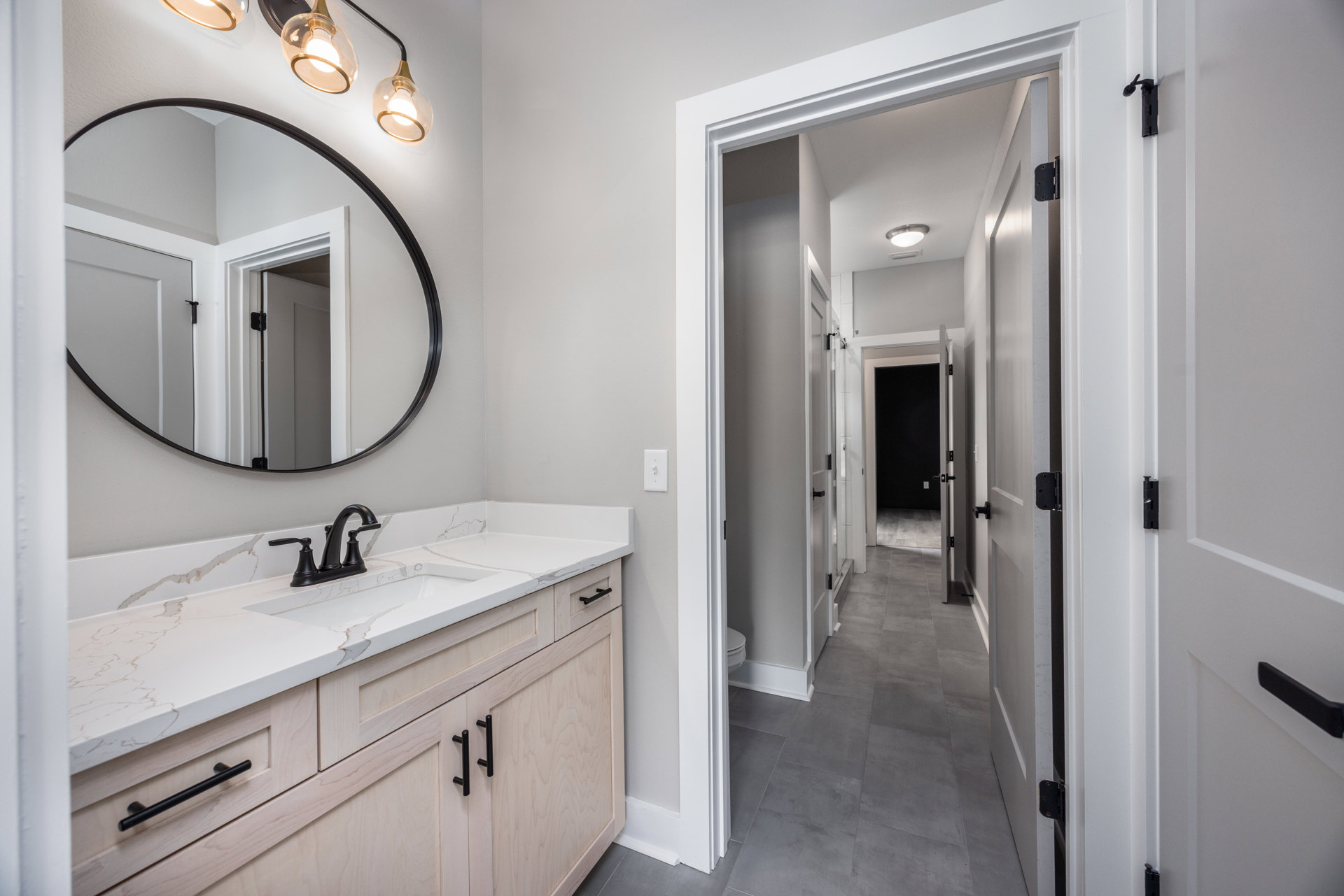 Bathroom with a round mirror above a white sink, black faucet, white countertop, light fixture with clear glass bulbs, tiled wall, and cabinetry with drawers.