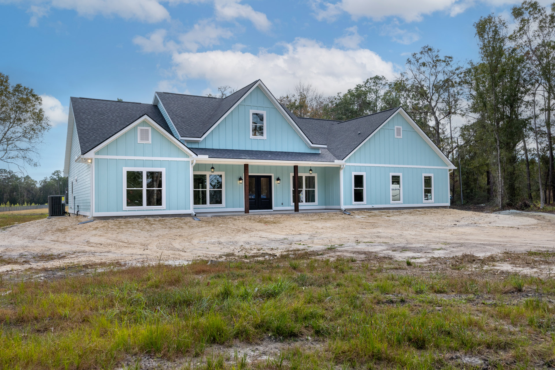 Blue cottage-style house with black doors and windows, one window showing broken glass, situated beside a dirt road and grassy field, surrounded by trees and cloudy sky; large