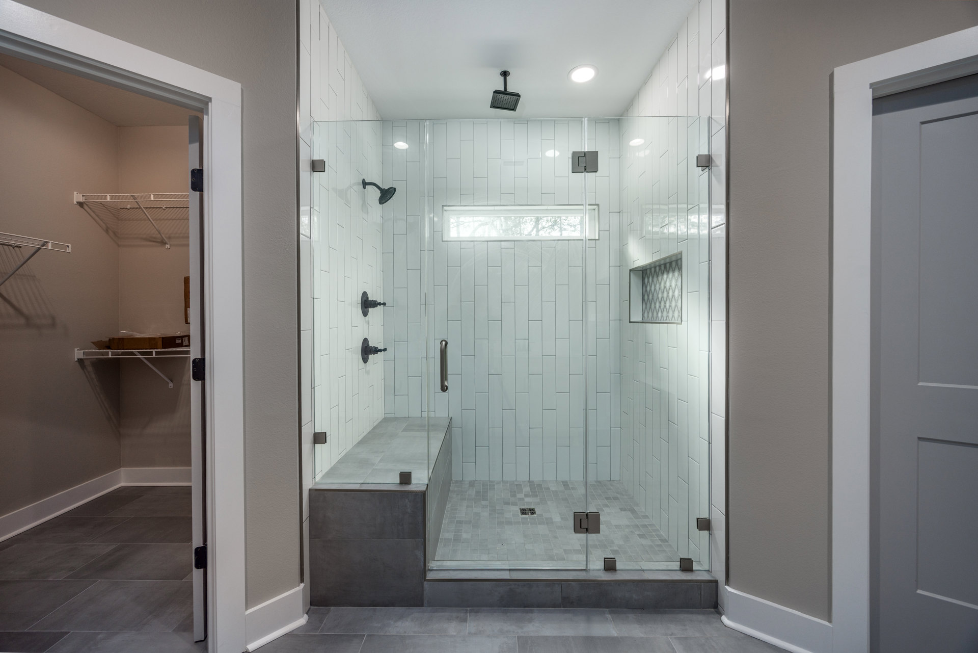 Glass shower enclosure with built-in bench, black shower head and pipe, white tile walls, glass railing along adjacent wall, window providing natural light.