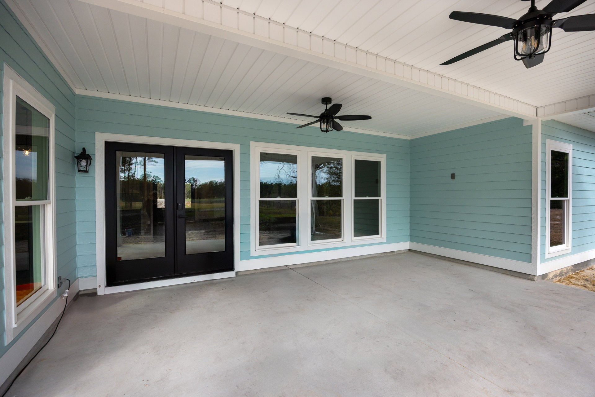 Living room with wood flooring, ceiling fan with light fixture, double glass doors opening to porch, white-framed windows, and trees visible outside.