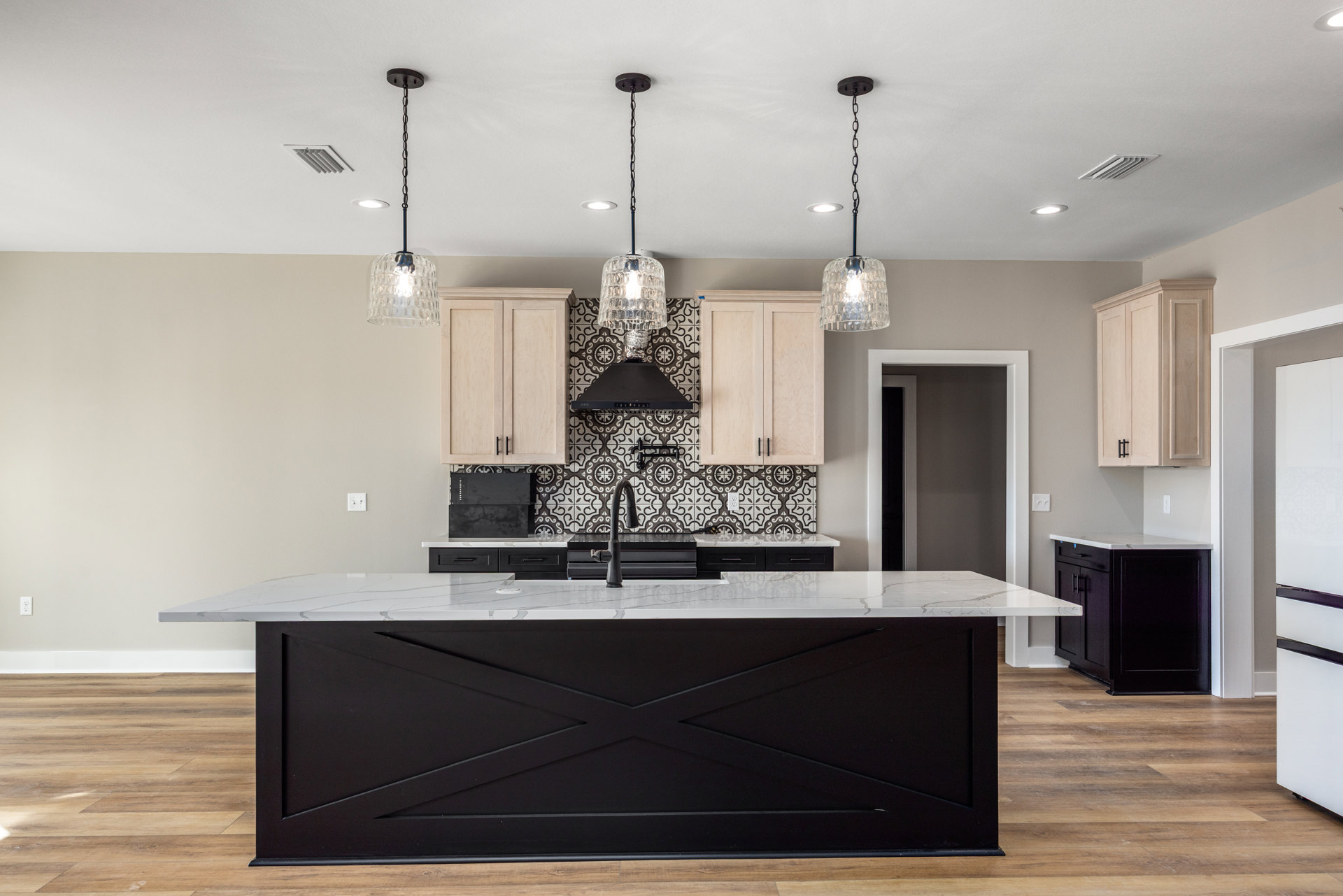 Spacious kitchen featuring a large white island with black cabinetry, black and white countertops, glass pendant light fixtures, and light wood flooring.