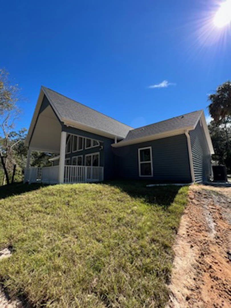 Two-story house with white-framed windows and a porch featuring white railings, set atop a grassy hill with trees and a dirt road in the foreground.