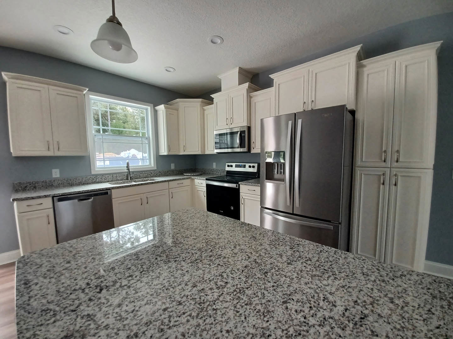 Granite countertops, stainless steel refrigerator, black and silver stove, glass door microwave, white framed window, wood cabinetry, and sink in a modern kitchen