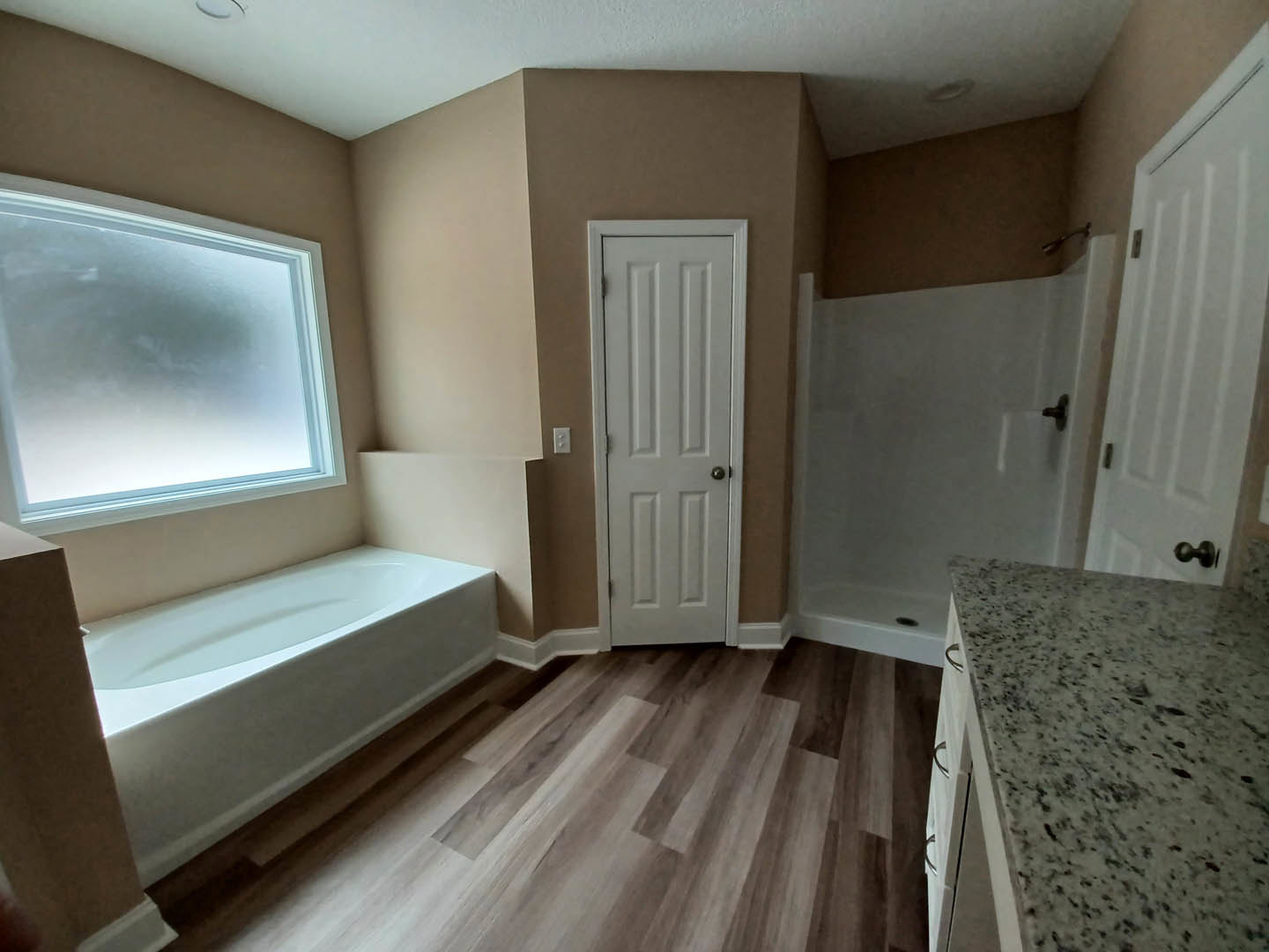 Bathroom with white bathtub and glass-enclosed shower, speckled black countertop, white door with silver knob, window above tub, light fixture on wall, neutral plaster walls and