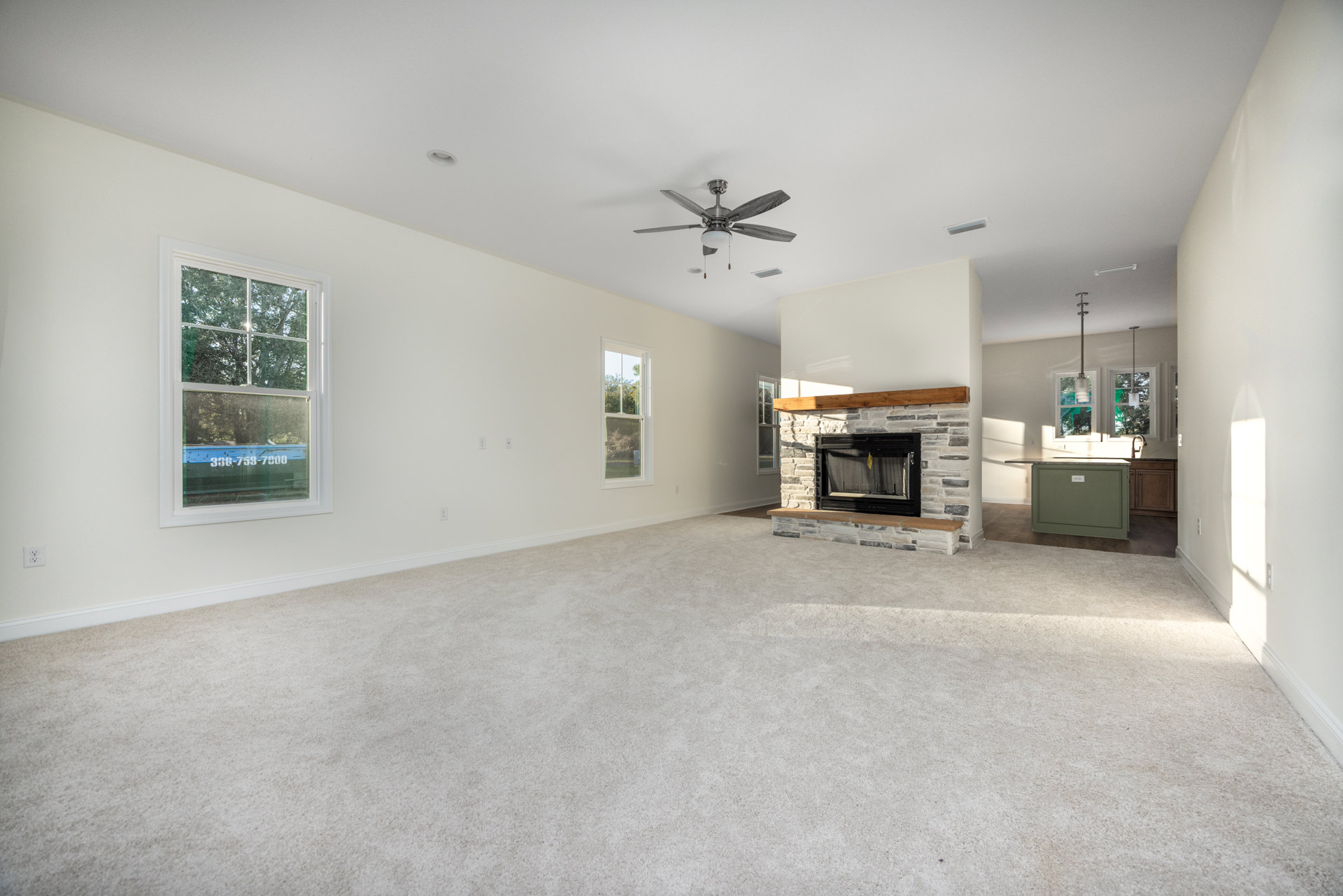 Carpeted living room featuring a stone fireplace with a wood beam mantel, ceiling fan with light, large window showing a blue box outside, and neutral plaster walls.