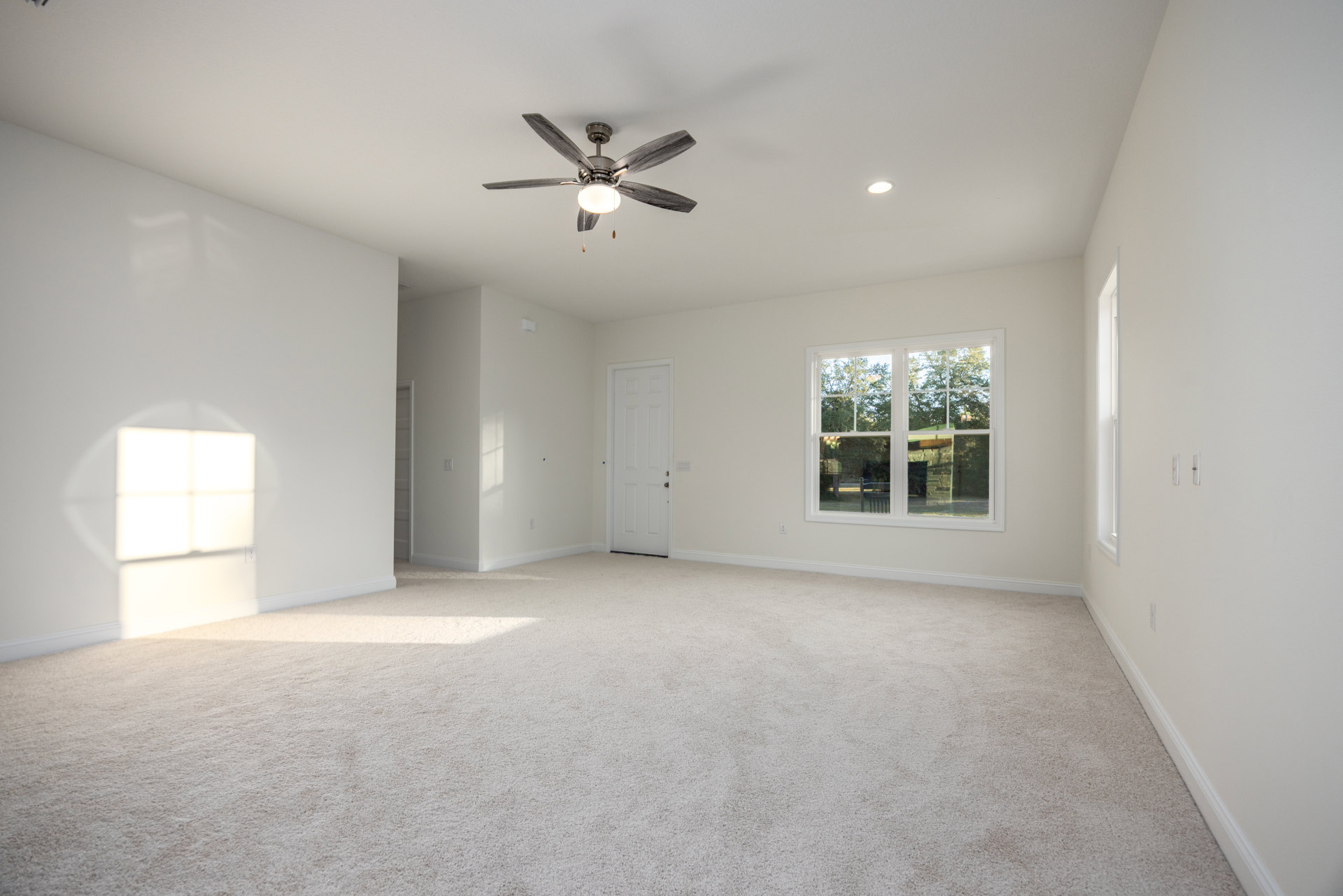 Bedroom with white walls, ceiling fan with light, single window showing green trees, white door with silver handle, light fixture on ceiling, neutral flooring