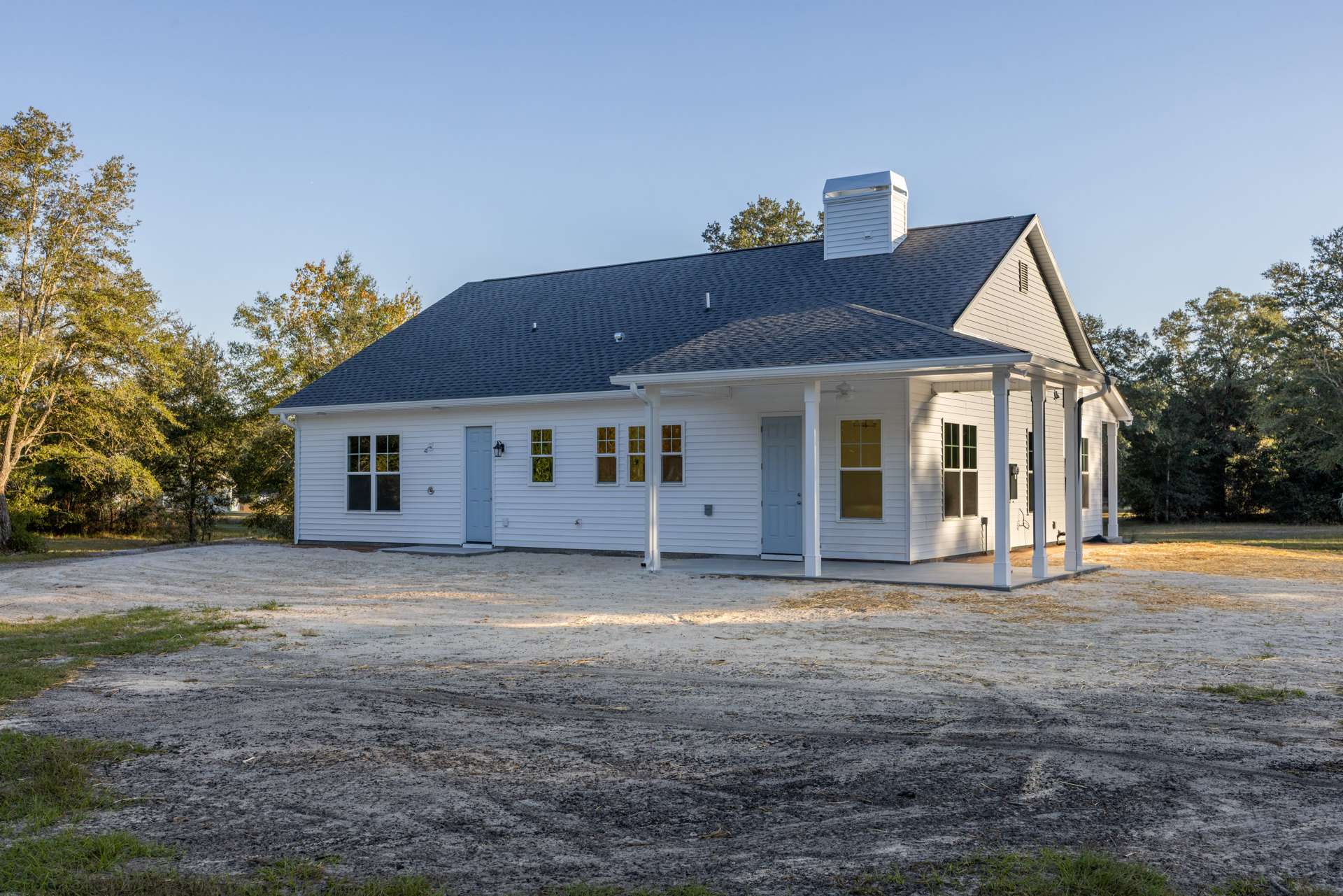 White siding house with blue metal roof, blue-framed windows and doors, leafy tree in front, dirt yard, white-framed window visible