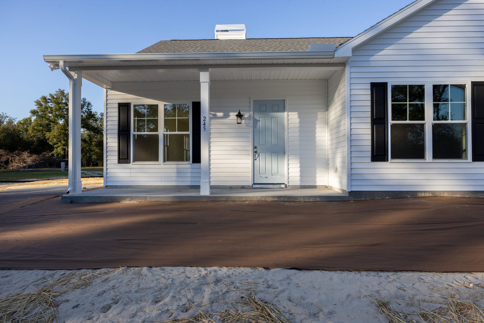 White siding house with covered front porch, white framed windows, blue door, and silver door handle; brown tarp and straw pile on sandy ground in foreground.
