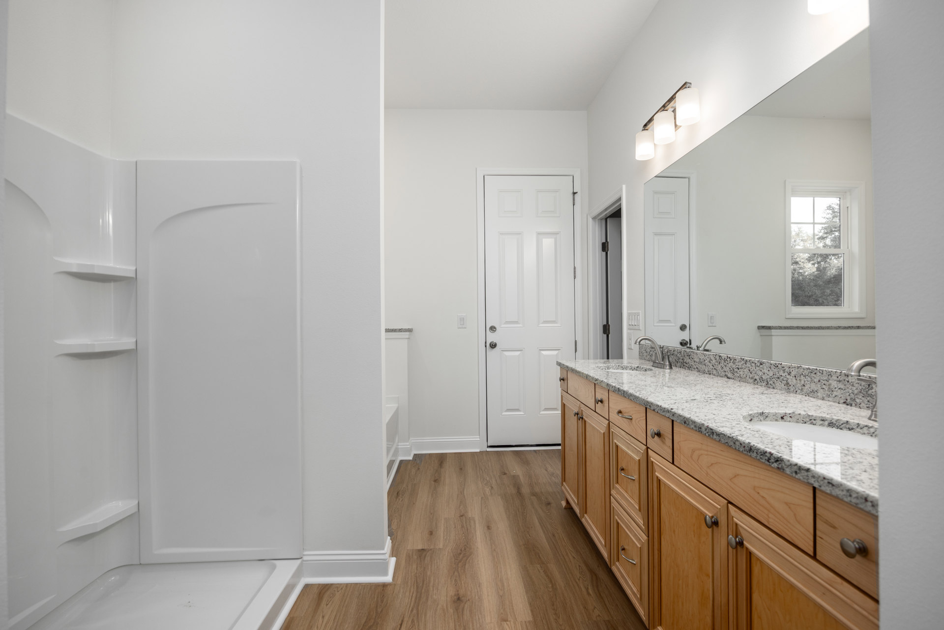 Bathroom featuring marble countertop, wooden cabinets, wood floor, white walls, silver-handled white door, window with outdoor trees, and rectangular white object on wall