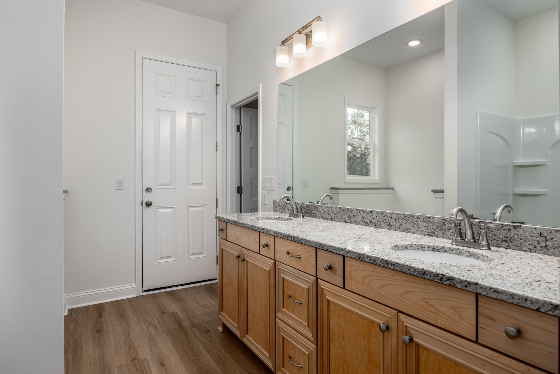 Bathroom with granite double sink vanity, large mirror, white cabinetry with silver knobs, tile flooring, window showing trees outside, modern light fixture, chrome faucet