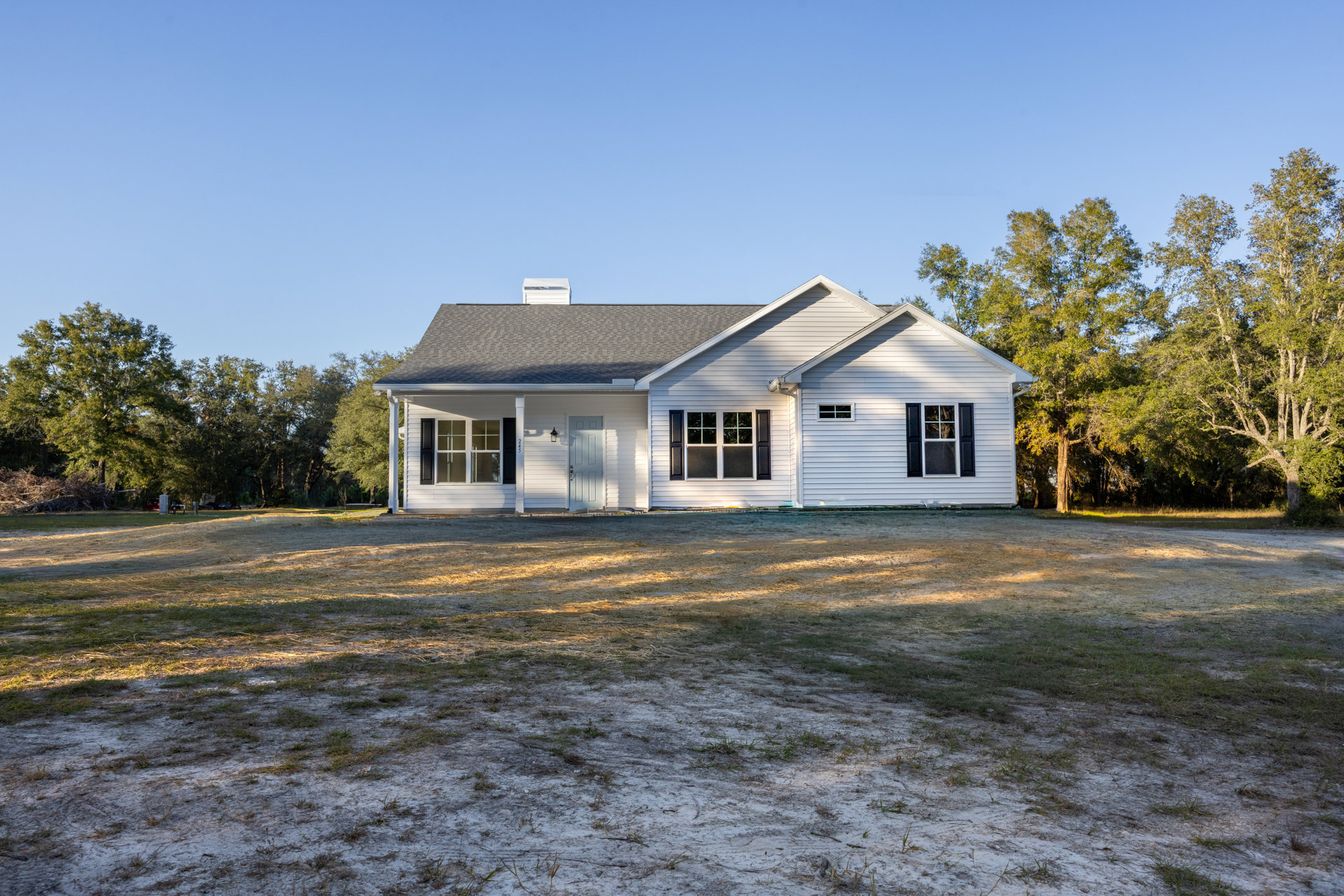 Two-story farmhouse with white door, white-framed windows, gray roof with chimney, surrounded by green lawn and mature trees