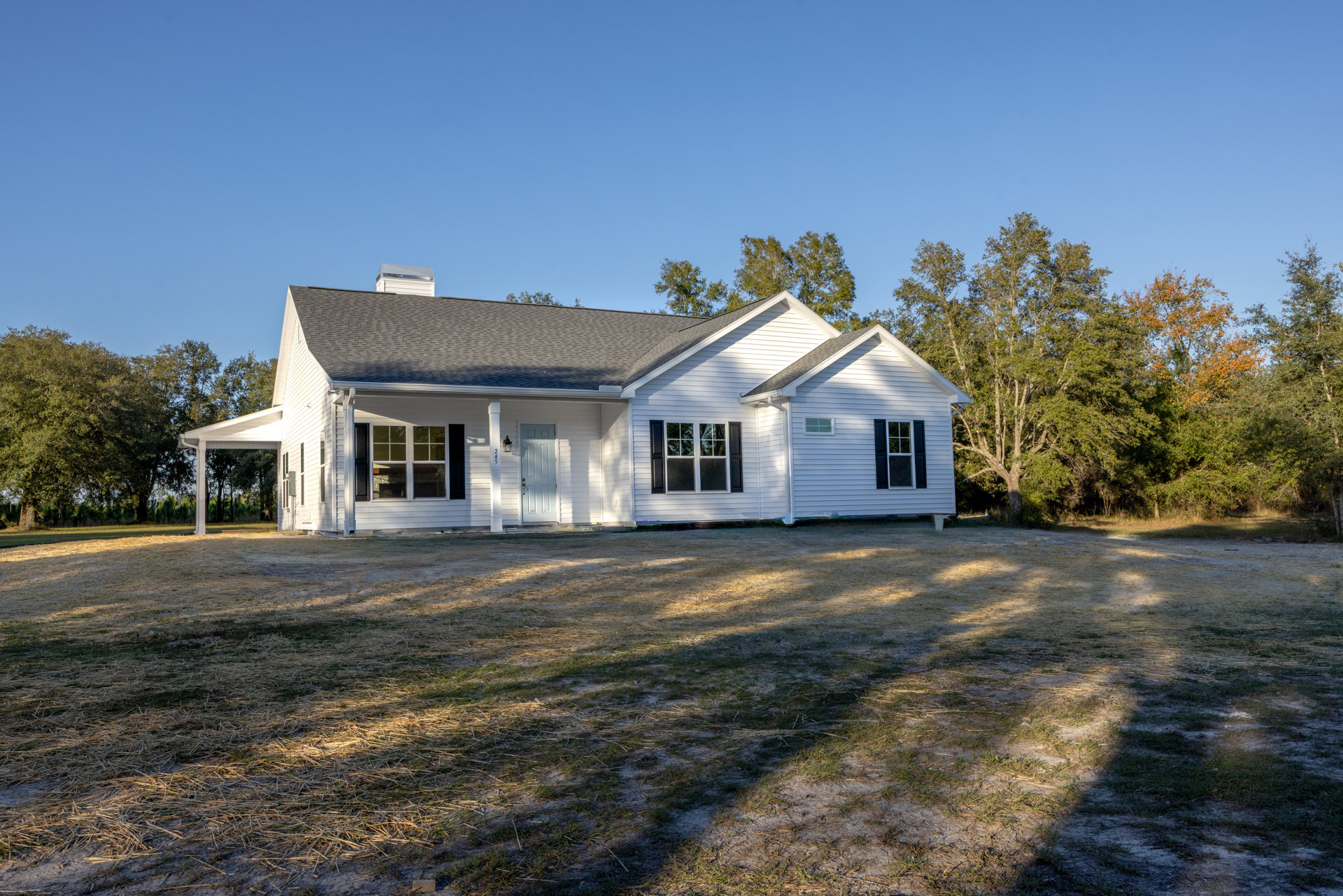 White siding farmhouse with black shuttered windows, white columned entry, white front door with side light, manicured lawn, mature trees behind, gabled roof