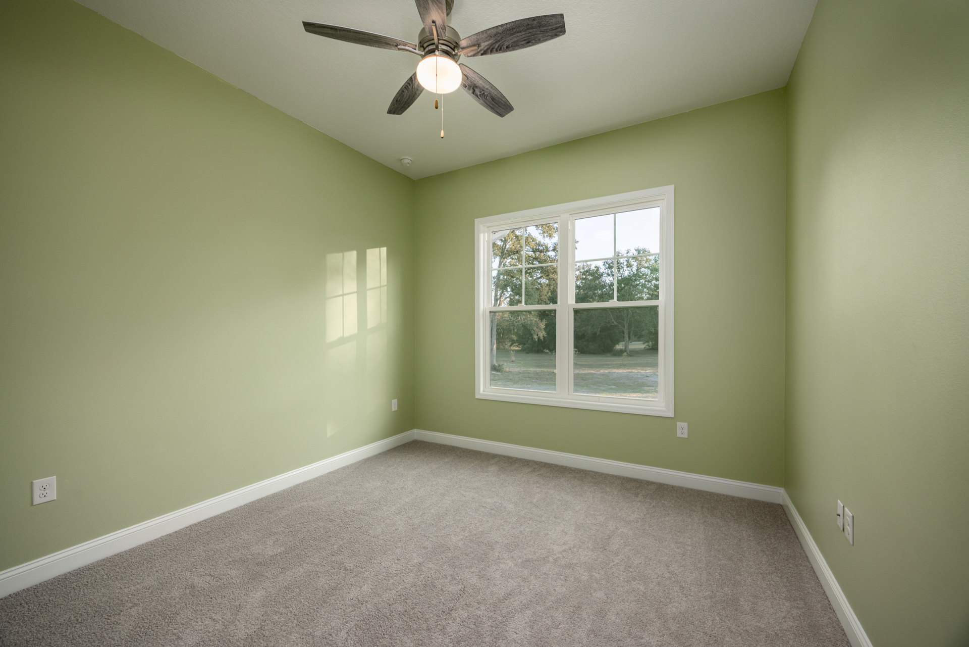 Neutral-toned carpeted room featuring a white ceiling fan with light fixture, single window overlooking greenery, smooth plaster walls, and minimal decor.