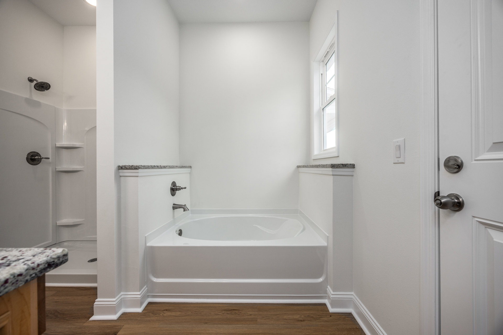 Freestanding white bathtub with chrome faucet beside wood flooring and tiled wall, bathroom vanity with countertop, window, and modern door knob visible