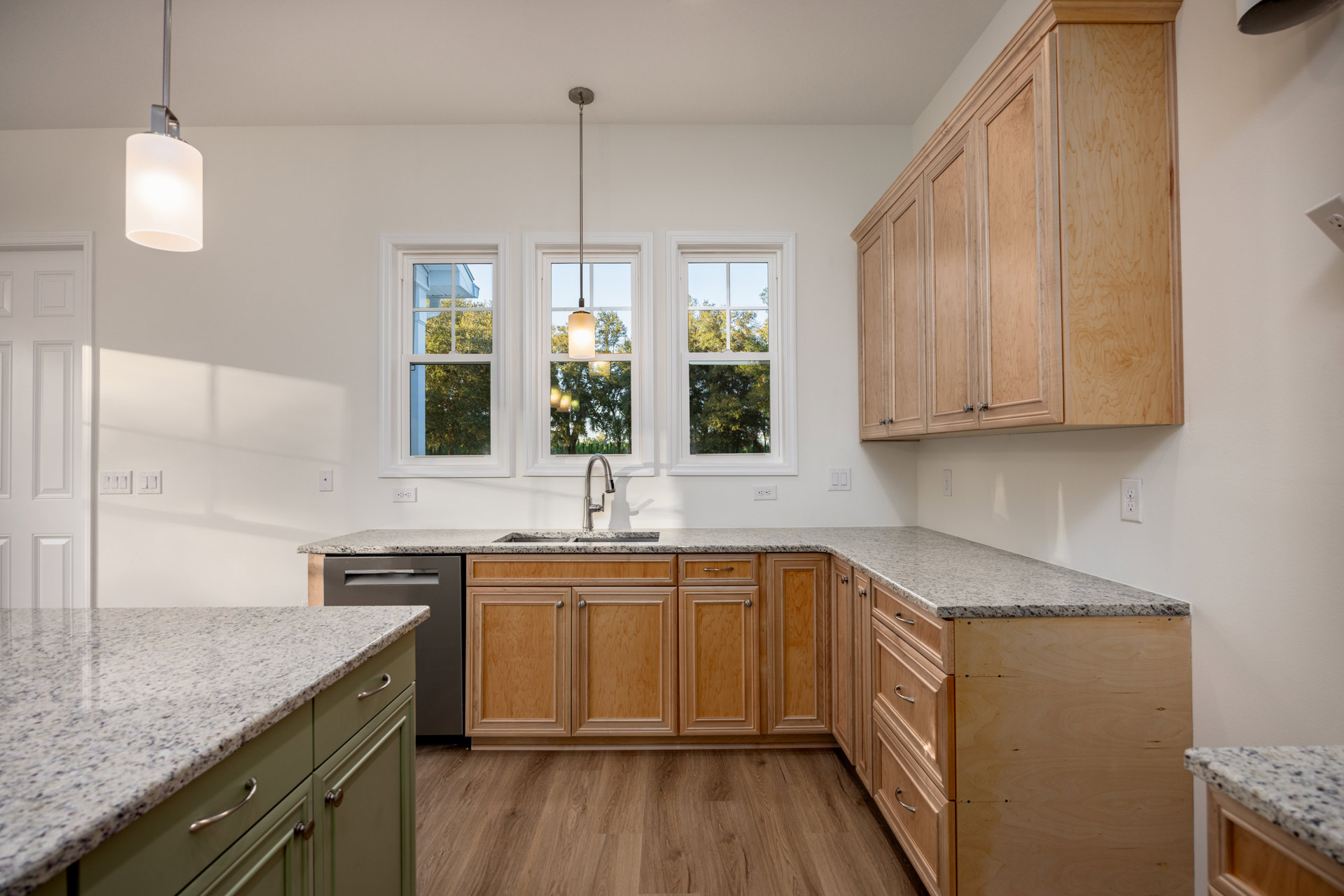 Kitchen featuring wooden cabinets, granite countertops, green lower cabinetry, stainless steel sink, ceiling light fixture, window overlooking trees, and wood door detail
