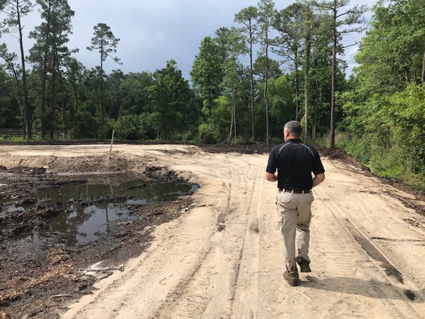 Man walking along a dirt road bordered by trees and plants under a clear sky, wearing casual clothing and hiking footwear