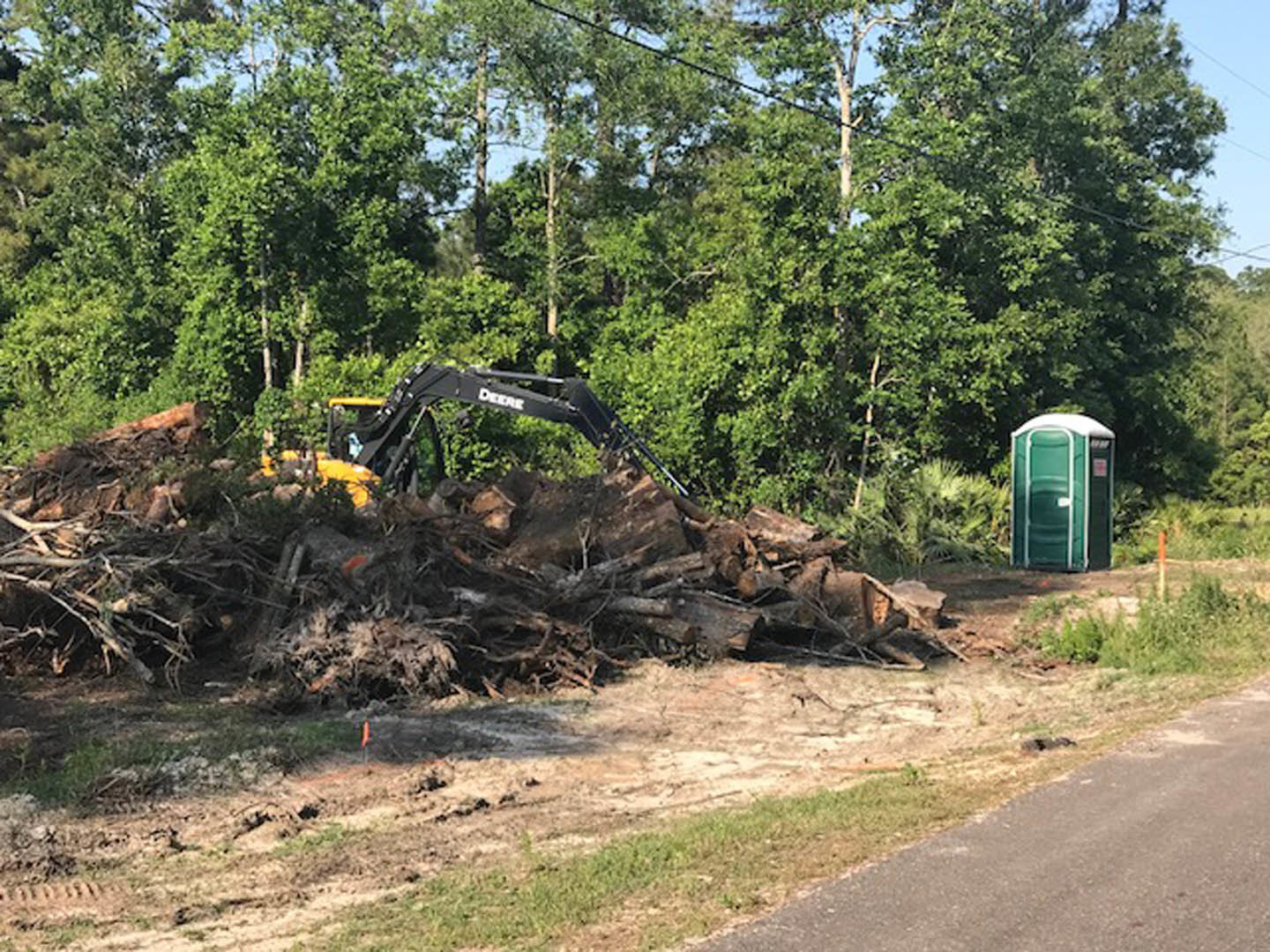 Green tractor parked on grassy yard beside a large pile of cut logs, surrounded by trees and open sky