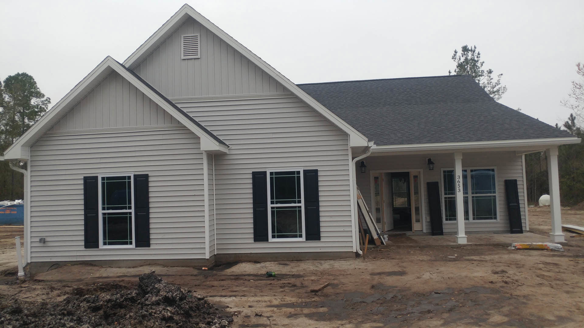 Partially built house with exposed siding, black shuttered window, dirt-filled yard, construction materials scattered, tree near roofline, clear sky overhead