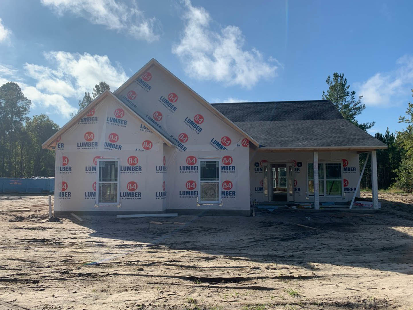 Framed house under construction with exposed plywood walls, white window frame, dirt lot in foreground, pitched roof, blue sky overhead