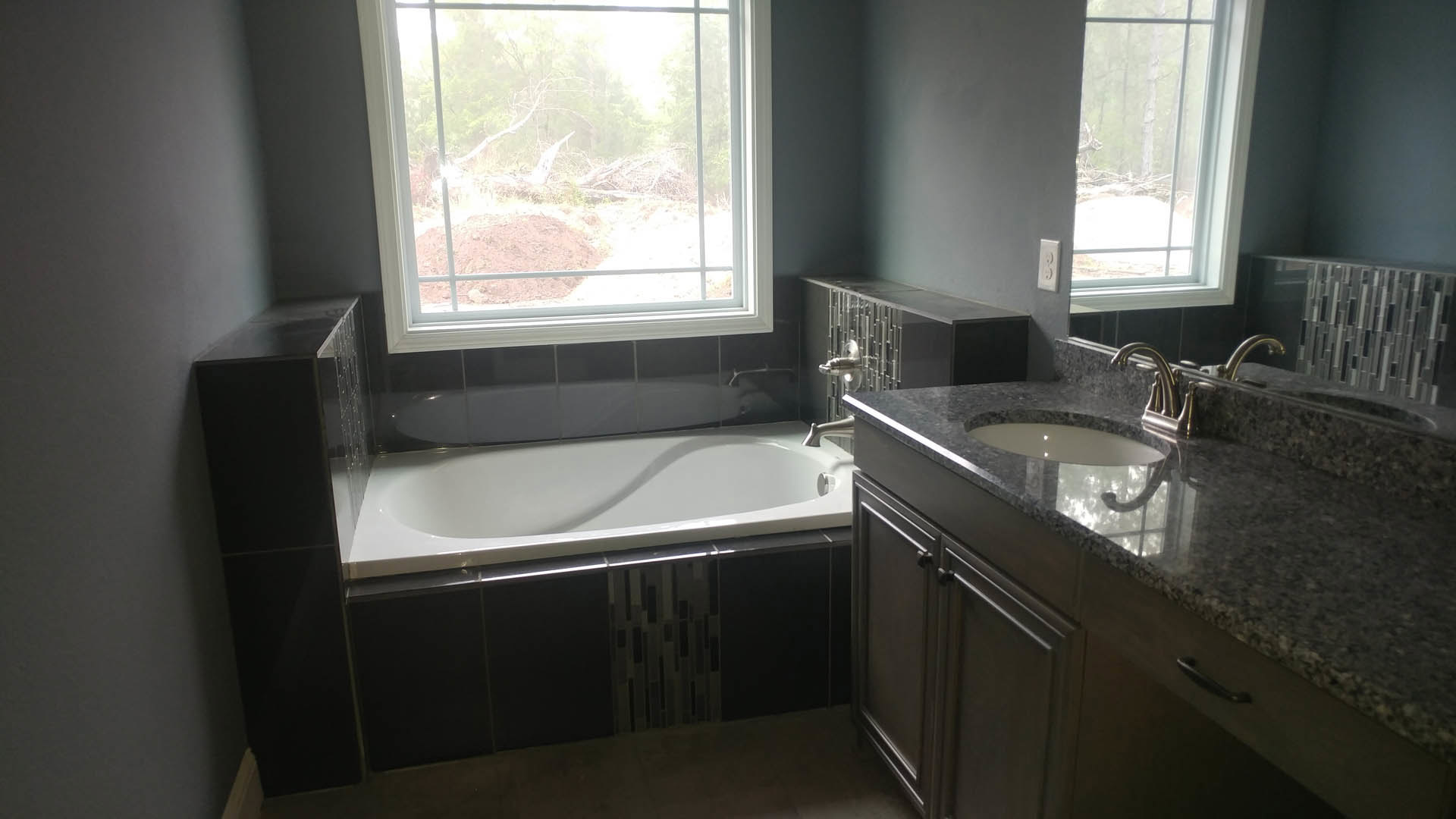 Freestanding bathtub and rectangular sink on stone countertop, large window overlooking dense forest, white tile walls, modern chrome fixtures
