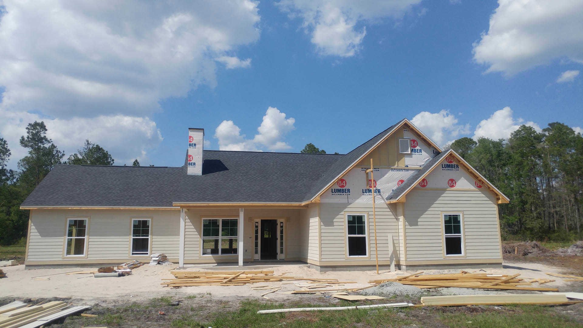 Partially built house with exposed framing, white window frames, black glass-paneled door, construction materials and wood pile on ground, window displaying a sign, blue sky and