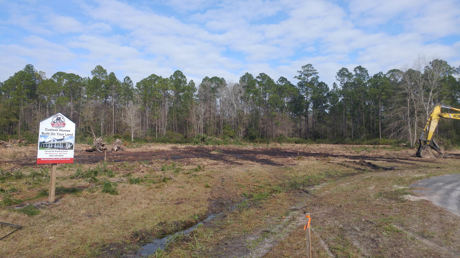 Muddy field with puddle, yellow excavator, house sign, grassy area, trees and blue sky with clouds in background