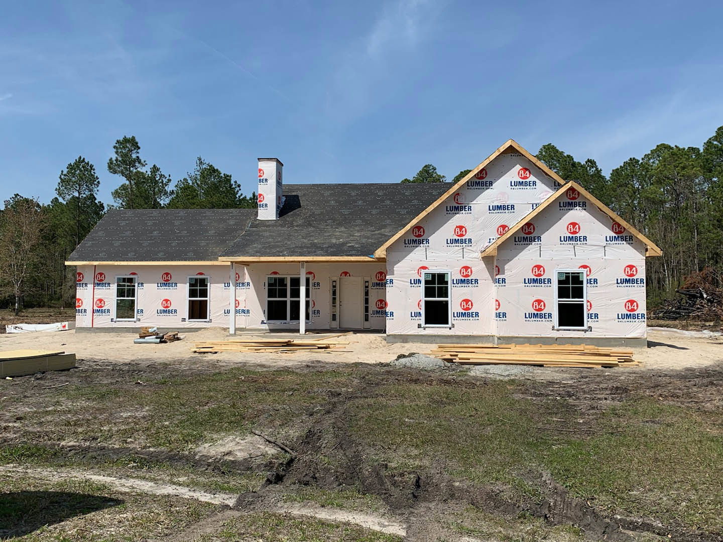 Partially built house with exposed framing, white window frames, plastic sheeting, chimney, and dirt yard, surrounded by trees under a cloudy sky