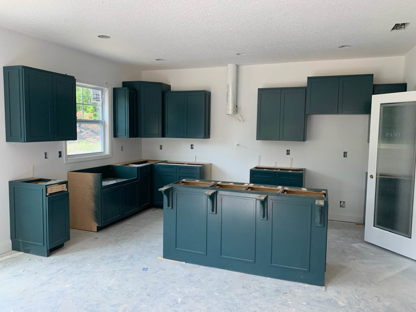 Green kitchen cabinets with brass hardware, white countertop, stainless steel sink beneath a window overlooking trees and mountains, light wood flooring