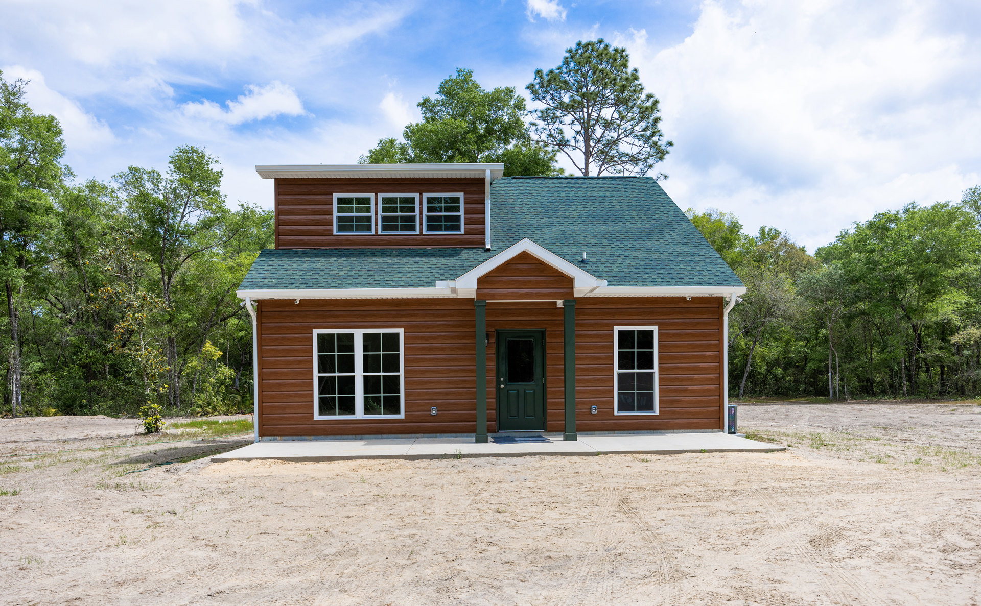 Two-story cottage with green front door, white-framed windows, green roof, gravel driveway, and dirt ground surrounded by trees under a cloudy sky