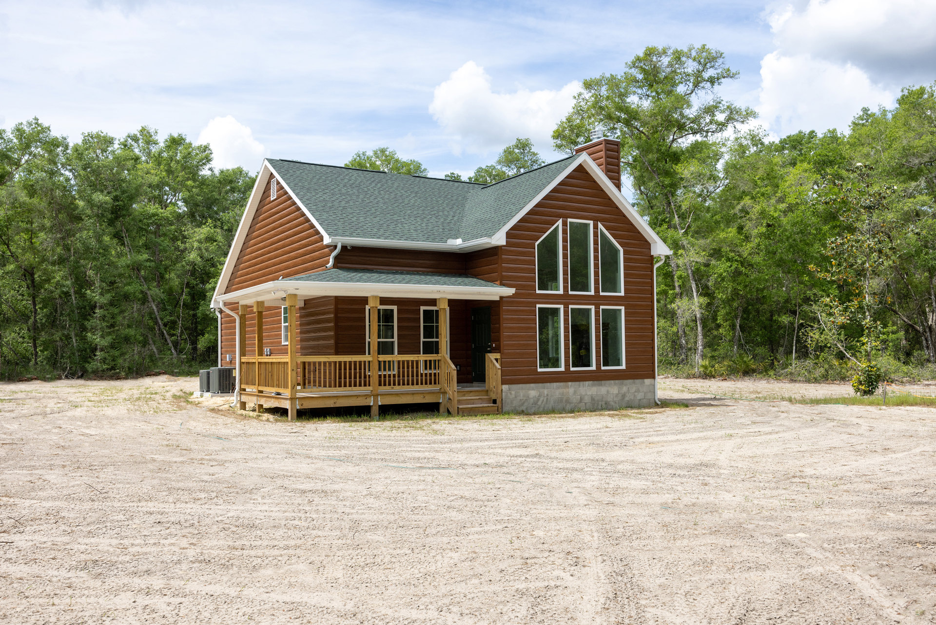 Wood-sided house with covered porch, wooden deck and railing, dirt lot in foreground, windows on facade, cloudy sky above