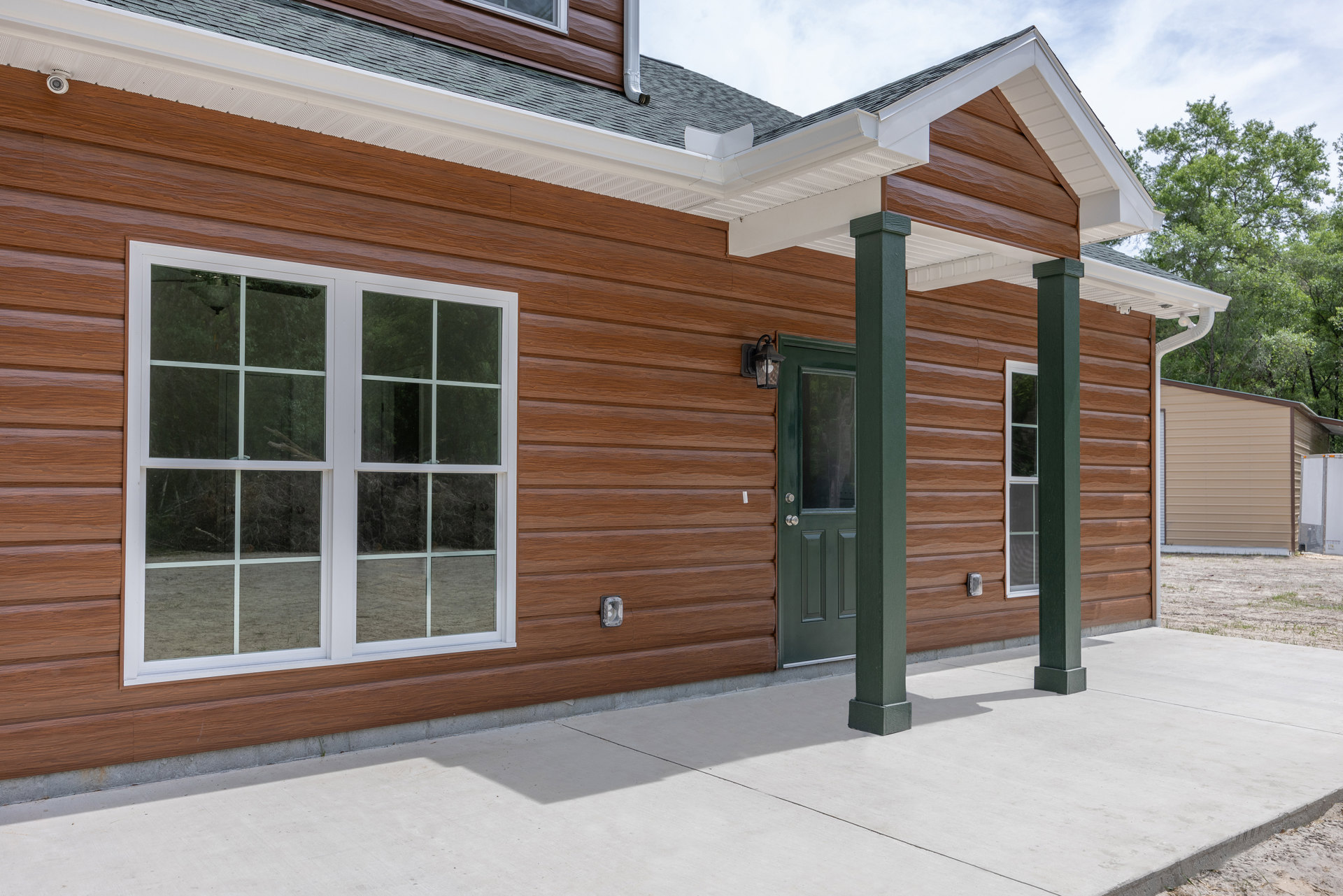 Front porch with concrete floor, green door, green column, white-framed window, light siding, and trees behind the house