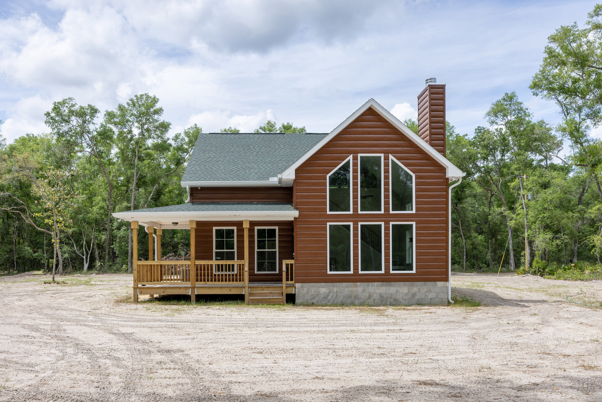 Two-story cottage with gray siding, white-trimmed windows, covered front porch, wooden deck with railing, brick chimney, surrounded by trees under a cloudy sky